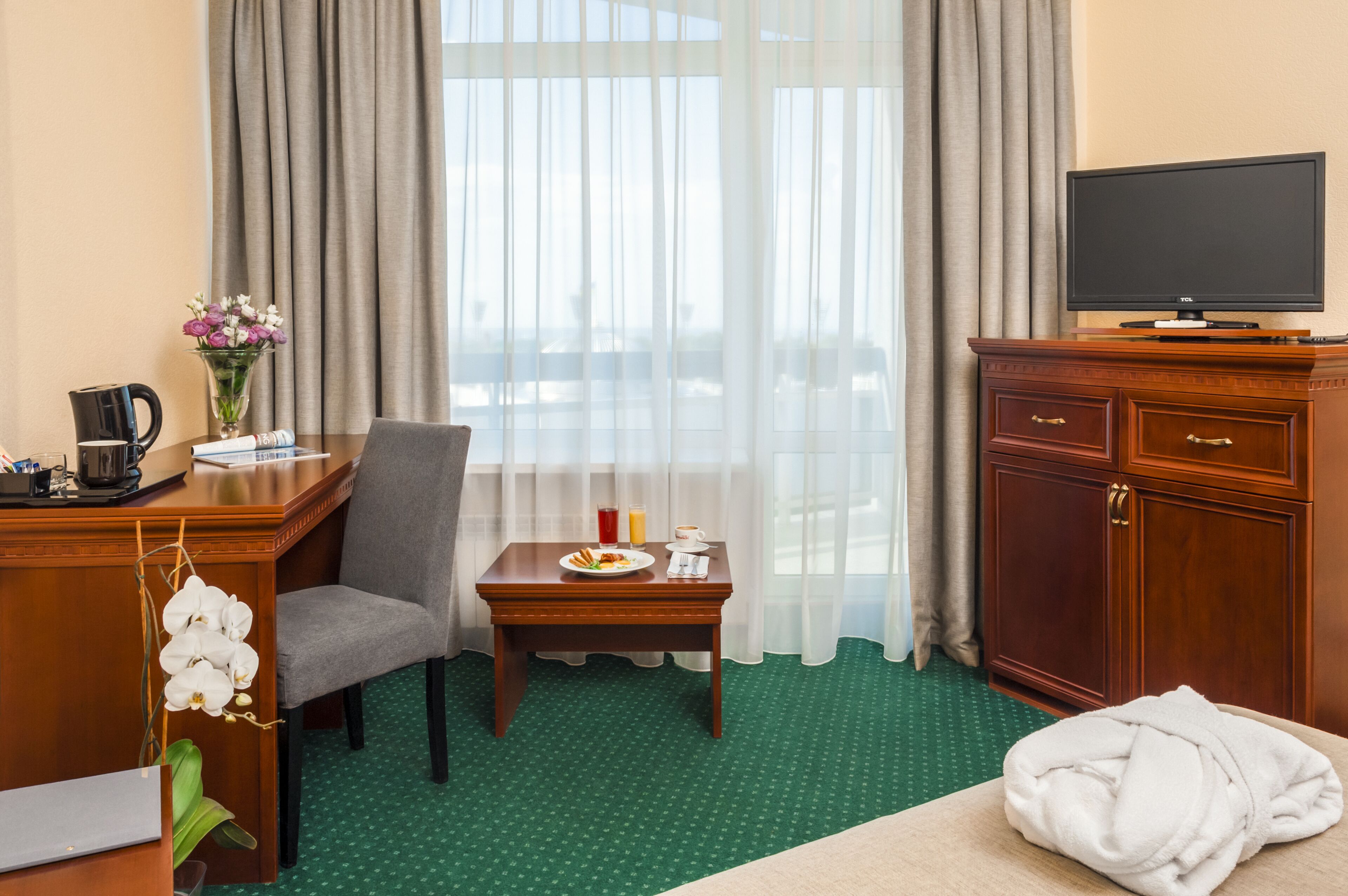 Une chambre d’hôtel comprenant un lit, un bureau avec une chaise, une petite table avec des articles pour le petit-déjeuner, un téléviseur posé sur un meuble et une fenêtre avec des rideaux.