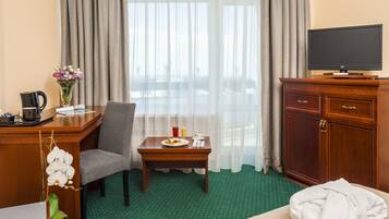Ein Hotelzimmer mit einem Bett, einem Schreibtisch mit Stuhl, einem kleinen Tisch mit Frühstücksangeboten, einem Fernseher auf einem Schrank und einem Fenster mit Vorhängen.