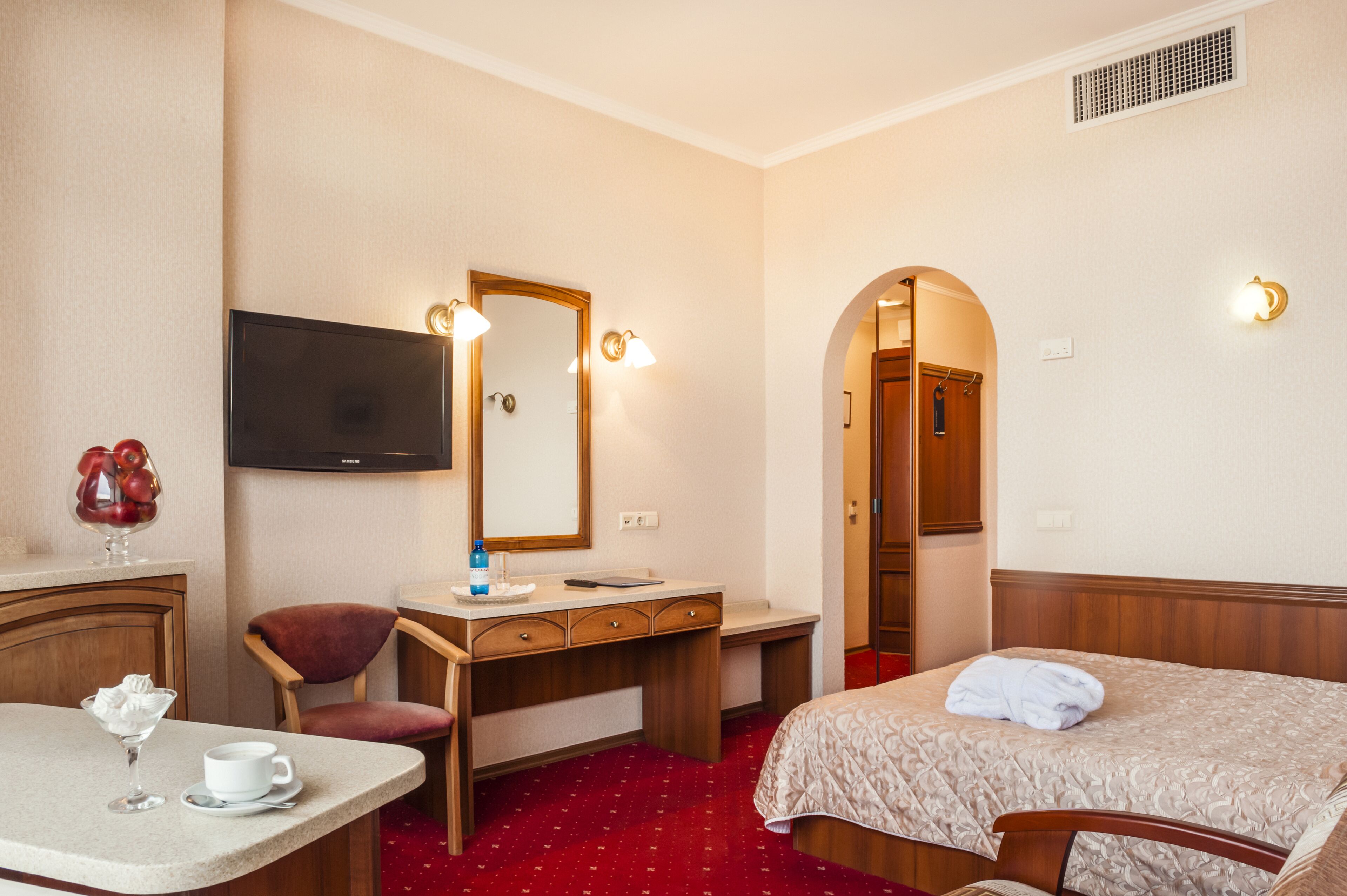 A hotel room with a bed, a desk with a mirror, a television, and a small table with a cup and a glass.