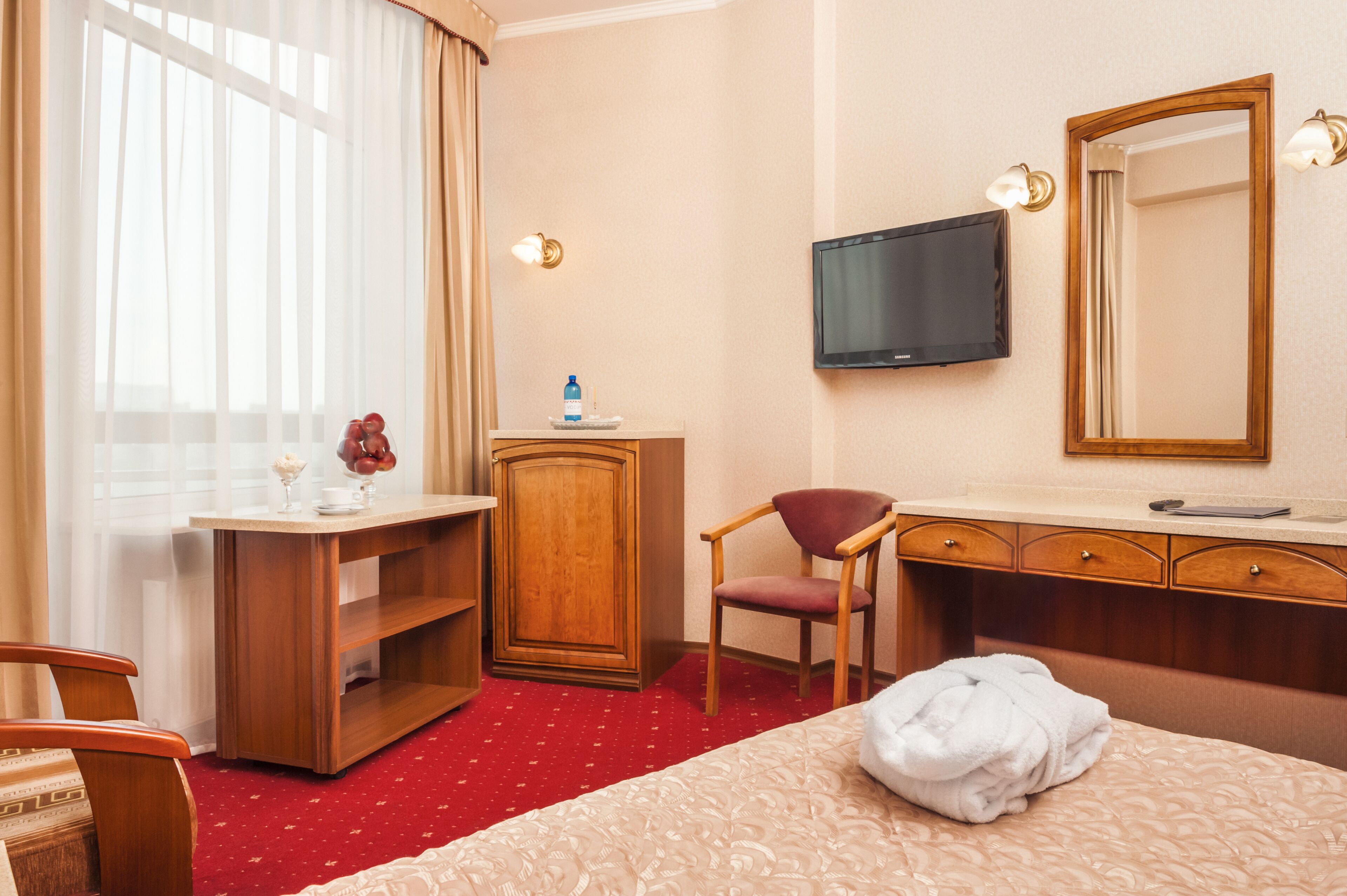 A hotel room with a bed, a desk with a chair, a television, a mirror, and a wardrobe.