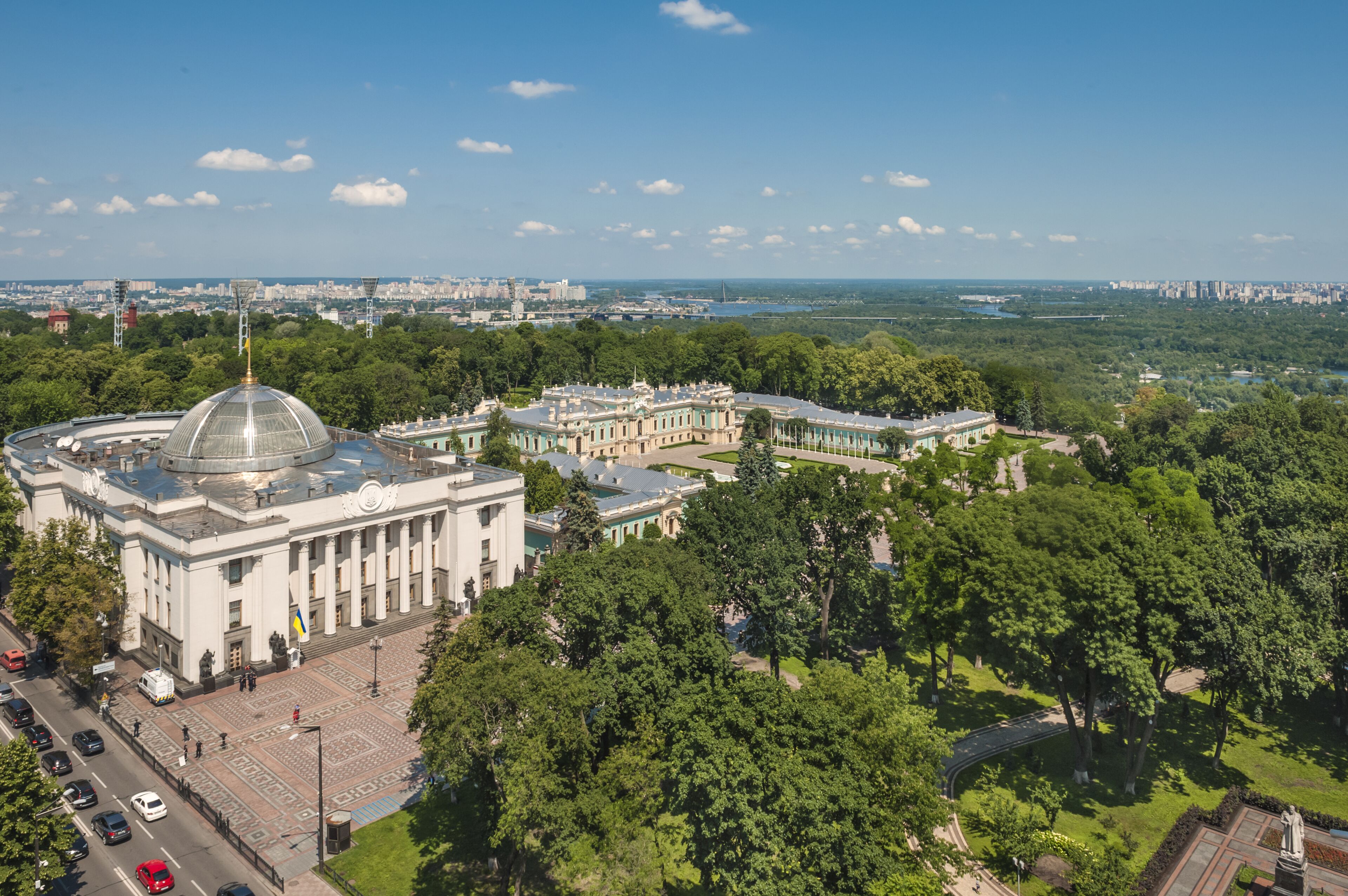 A large, historic building with a central dome and classical architecture, surrounded by lush green trees and a clear sky.