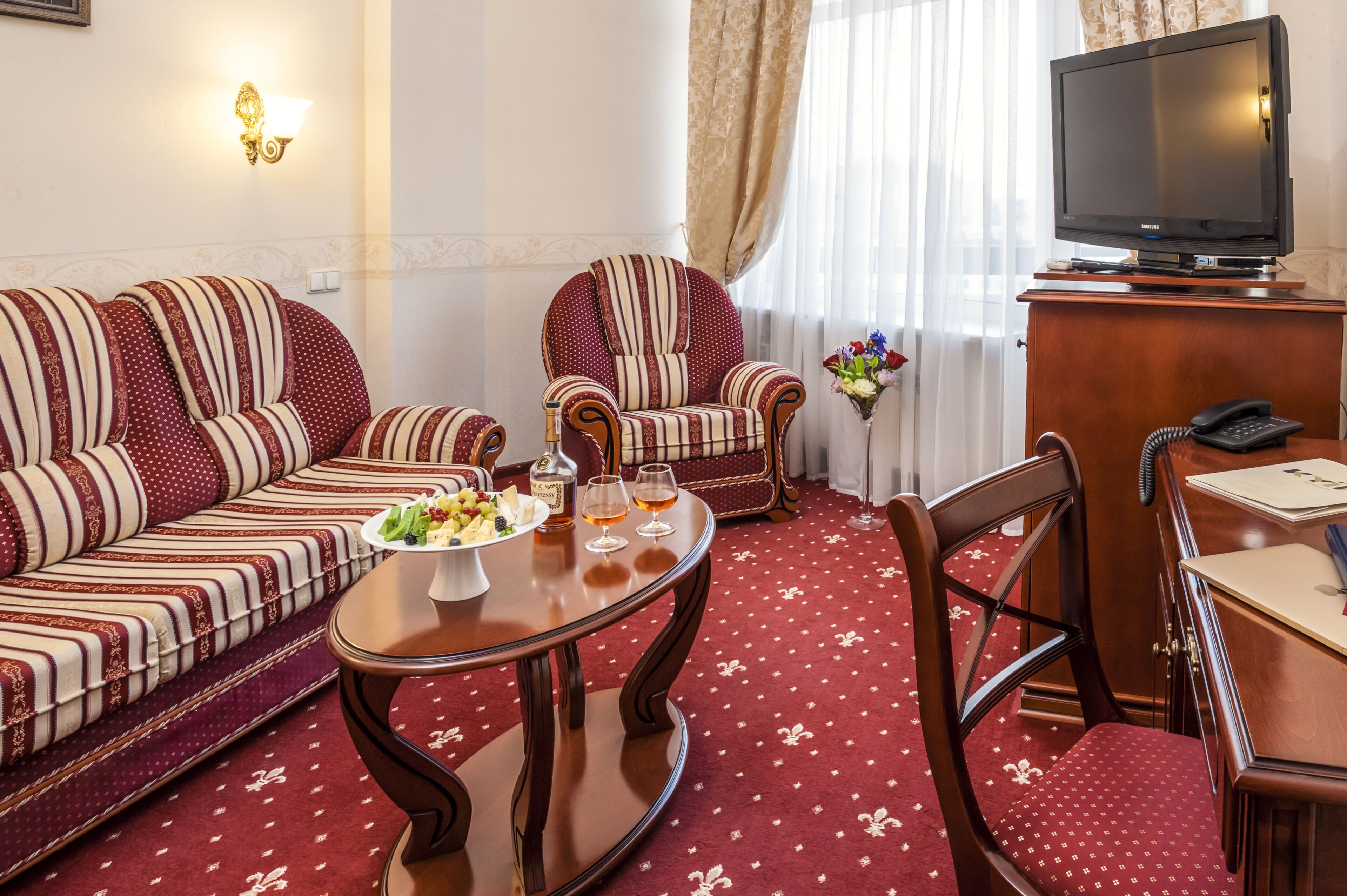 A hotel room with a sofa set, a dining table with a fruit platter, a chair, a television, and a desk with a telephone.