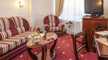 A hotel room with a sofa set, a dining table with a fruit platter, a chair, a television, and a desk with a telephone.