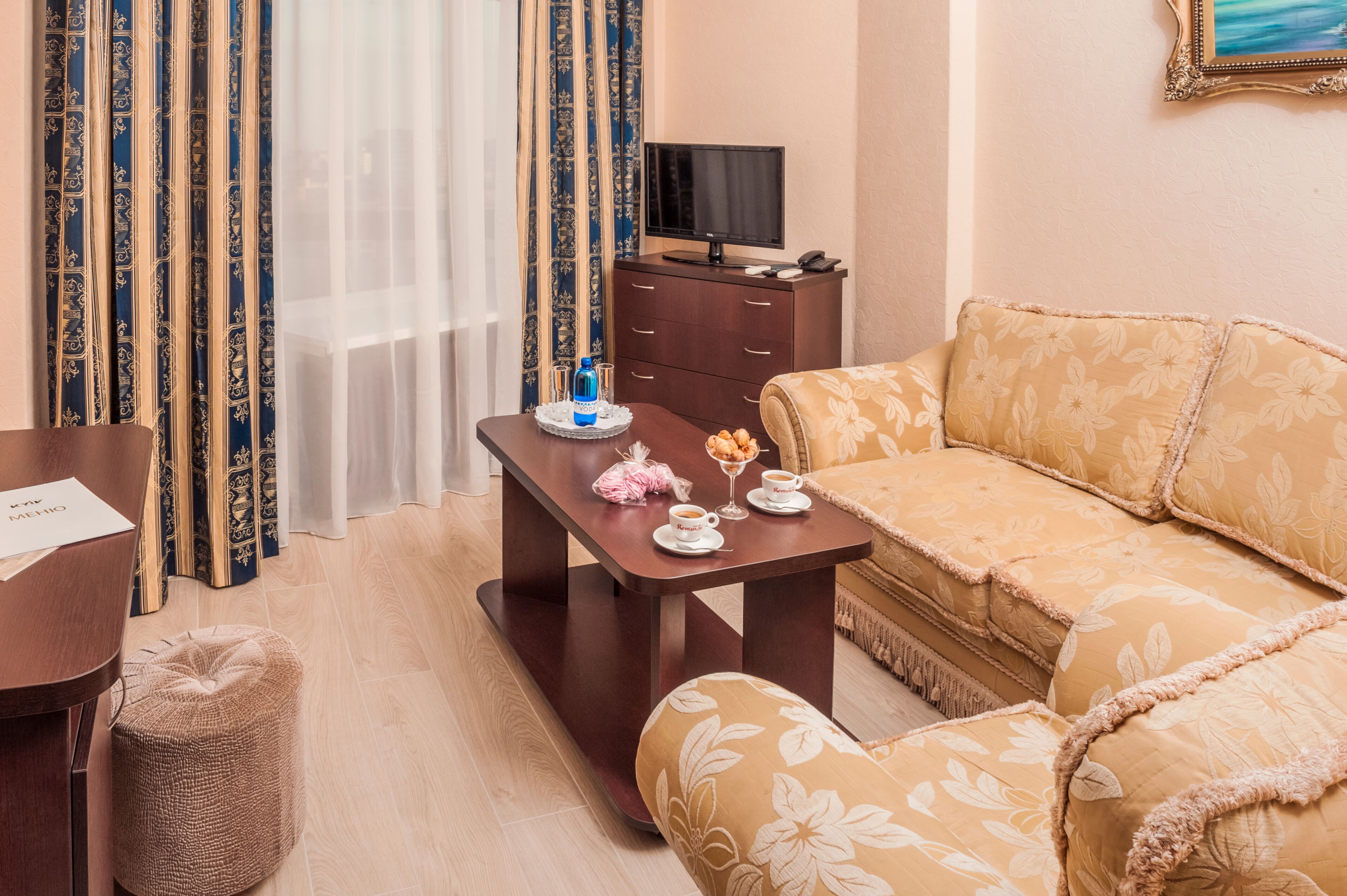 A hotel room with a sofa, a wooden table with a tray of snacks, a television, and a curtain with a floral pattern.