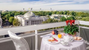 Ein Balkon mit Frühstückstisch, mit Croissants, Obst und Tee, mit Blick auf eine Stadtlandschaft mit einem markanten Gebäude und Grünflächen.
