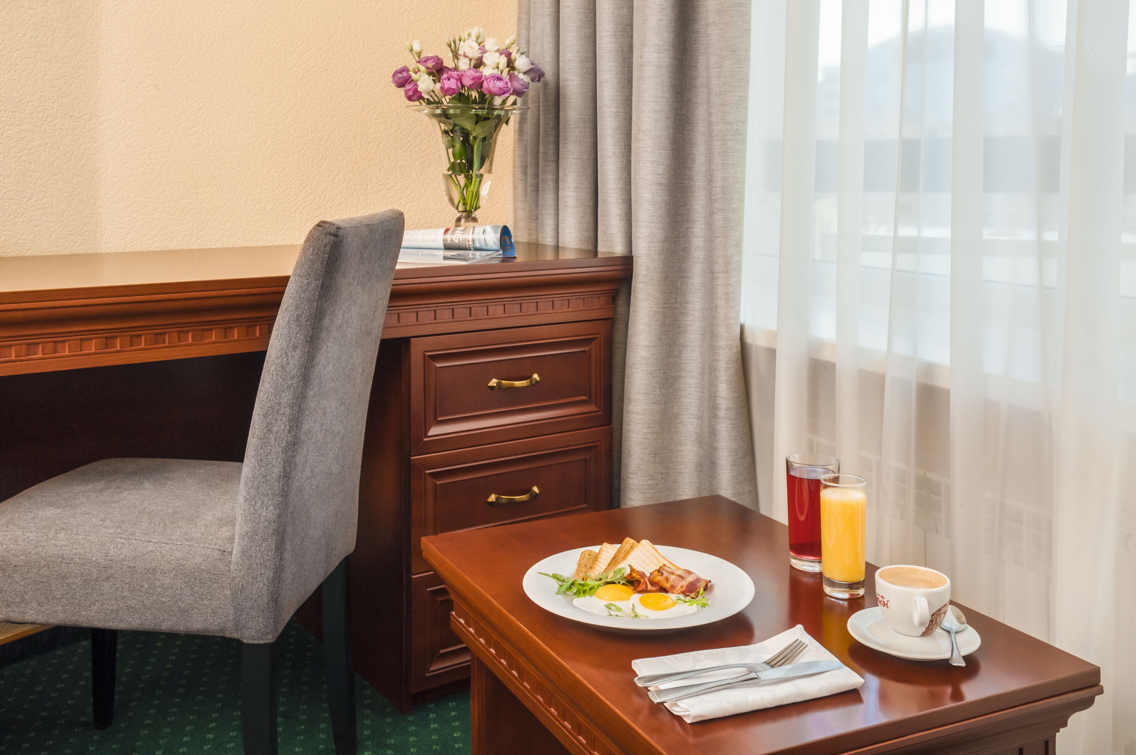 A hotel room with a wooden desk, a grey chair, a vase with flowers, a plate with breakfast, a glass of orange juice, and a cup of coffee.