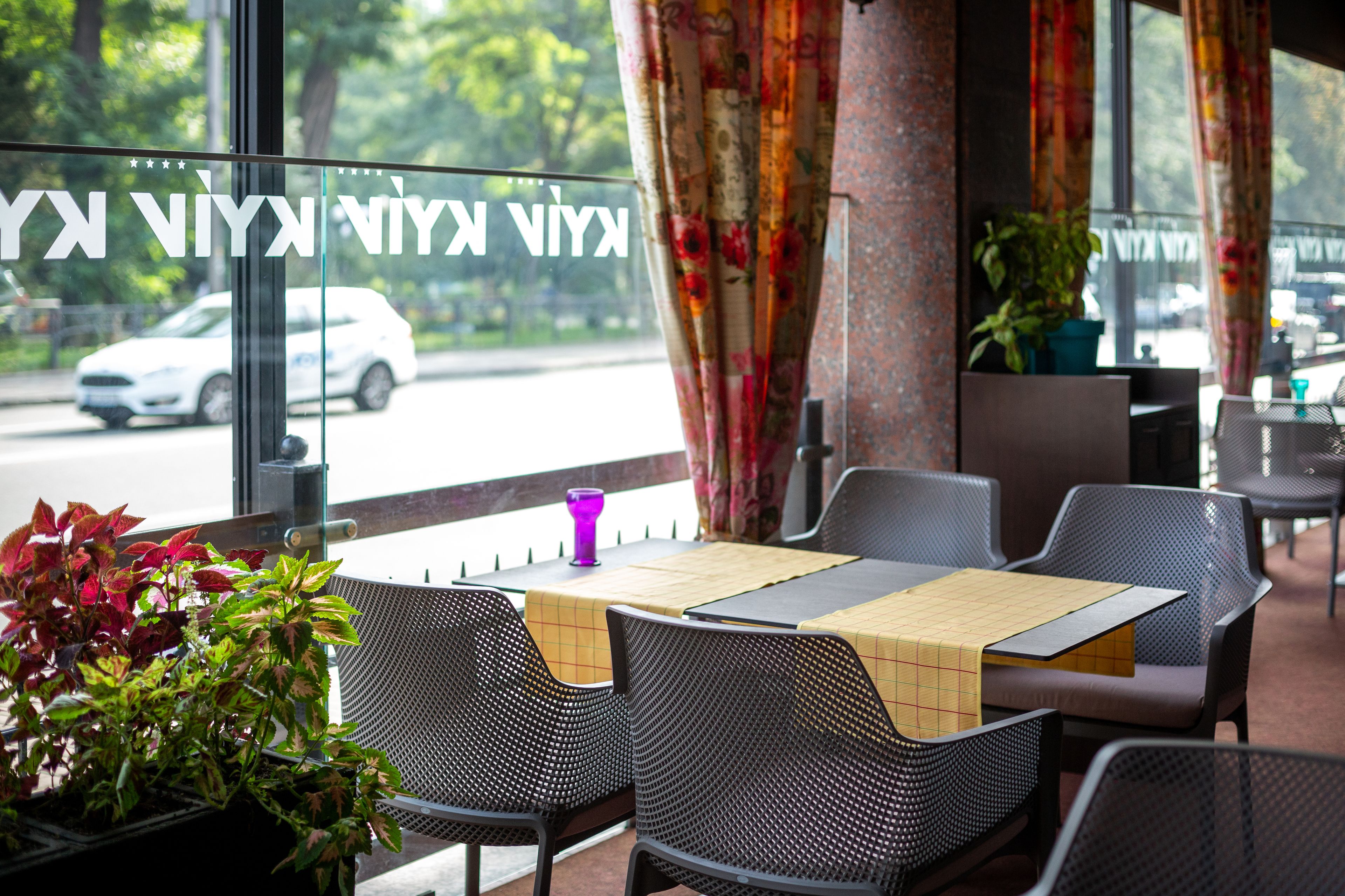 An outdoor seating area with wicker chairs and a table, a potted plant, and a view of the street through a window with curtains.