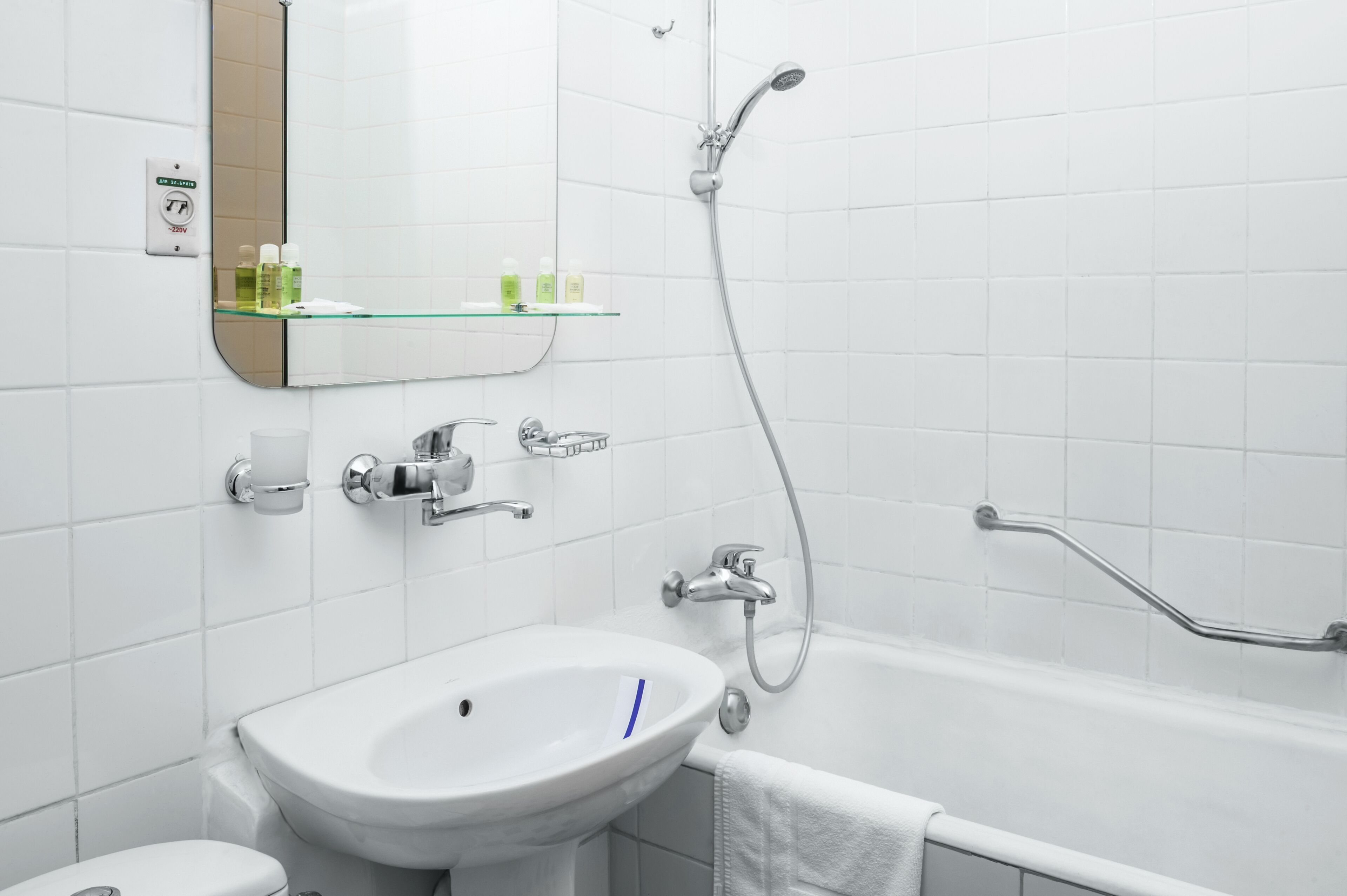 Een badkamer met een wit wastafelmeubel, een spiegel, een glazen plank met toiletartikelen, een handdouche en een handgreep in de douche.