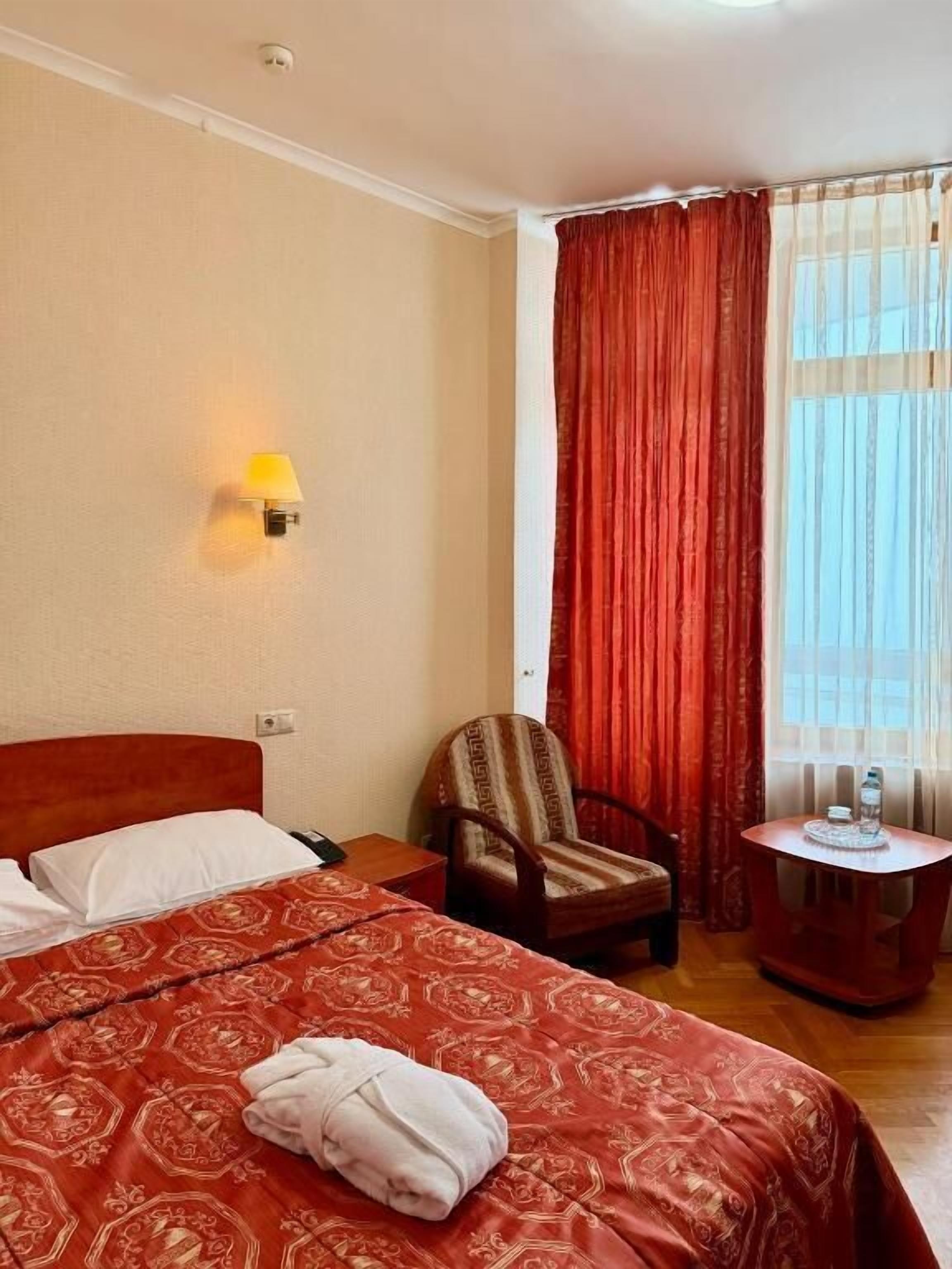 A hotel room with a large bed, a chair, a small table with water bottles, and a window with curtains.