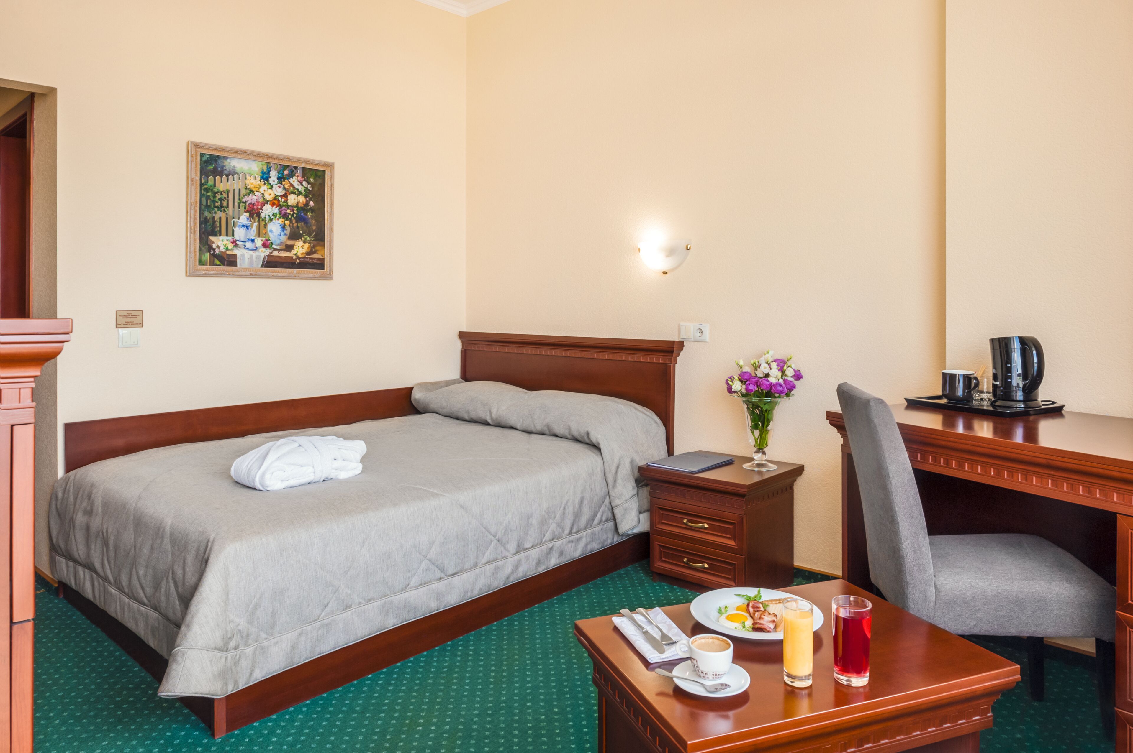 A hotel room with a bed, a desk, a chair, a coffee maker, a vase with flowers, a plate with food and a drink, and a painting on the wall.