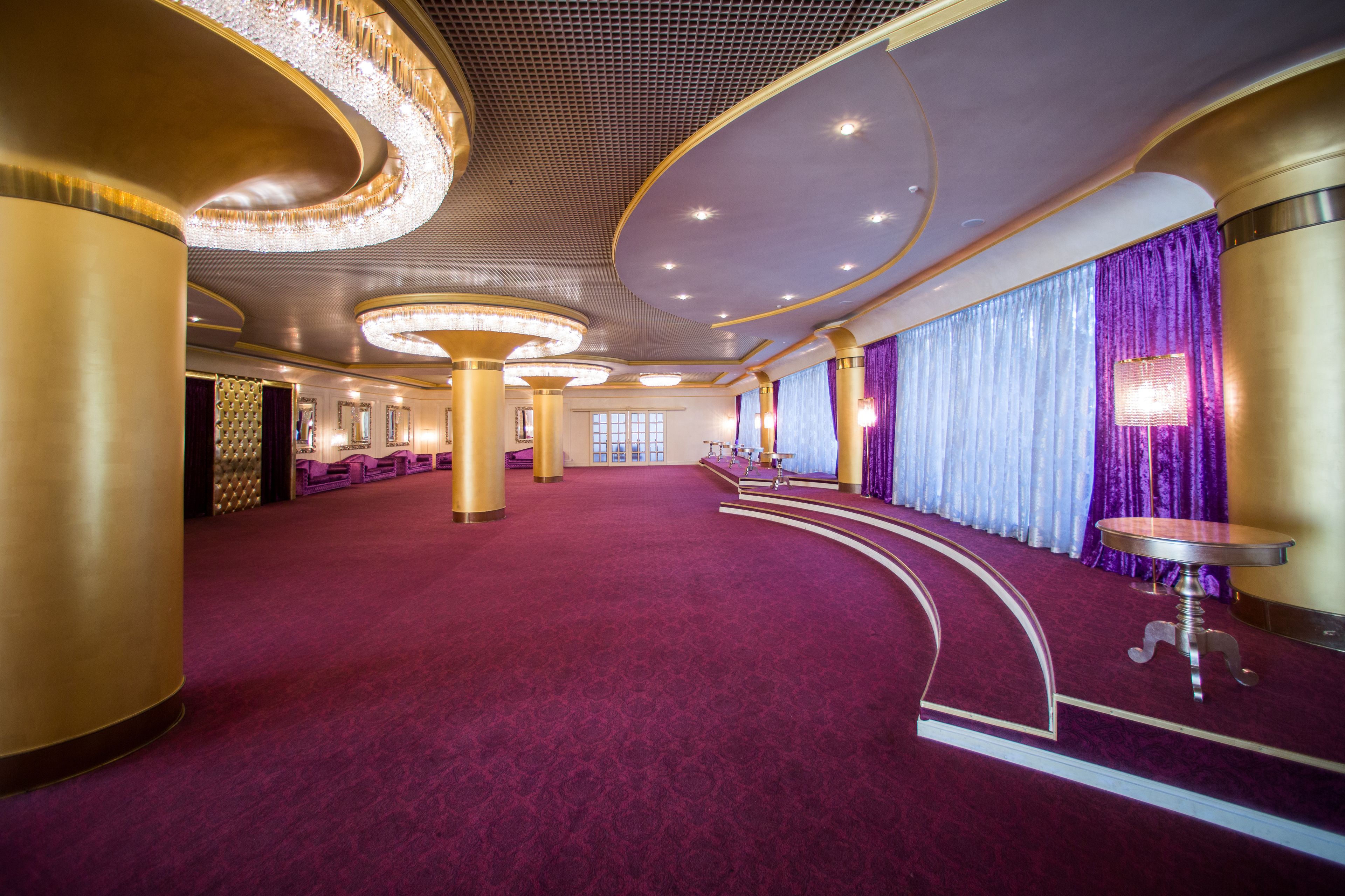 A spacious, elegantly decorated hall with a red carpet, large columns, and a stage area.