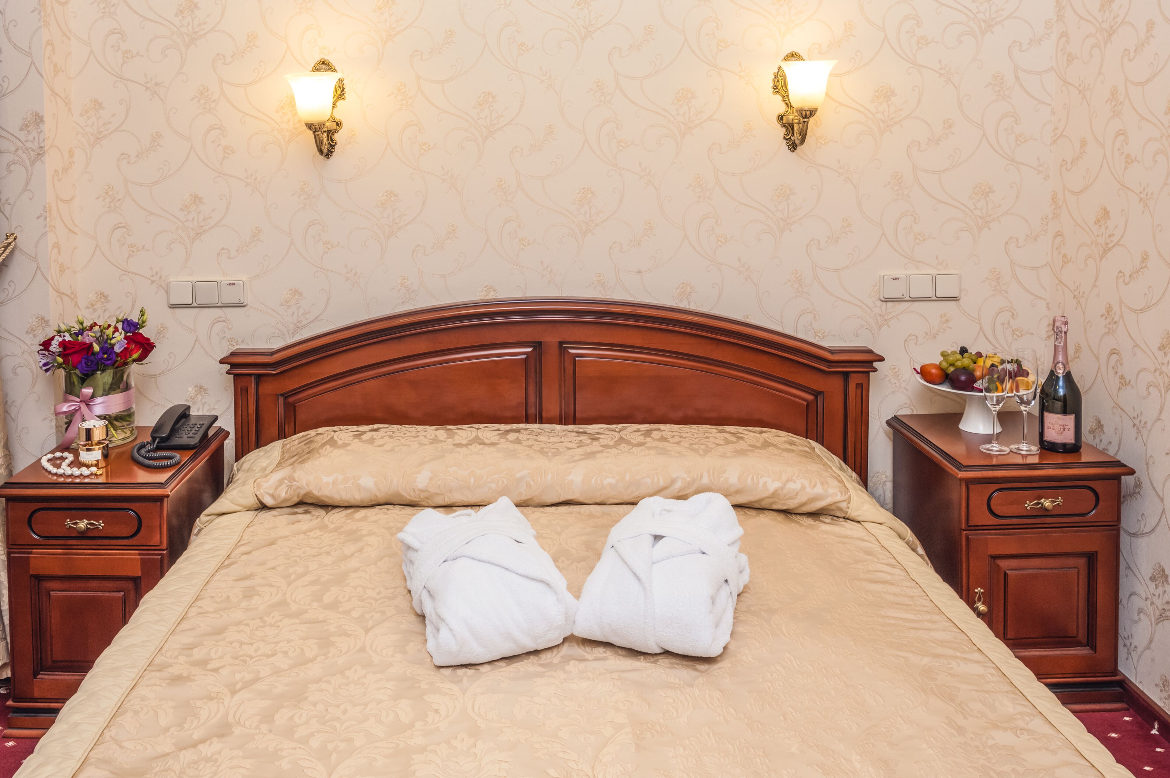 Een netjes opgemaakt bed met twee handdoeken, een nachtkastje met een telefoon en een bloemenvaas, en een bijzettafeltje met een fles en glazen.