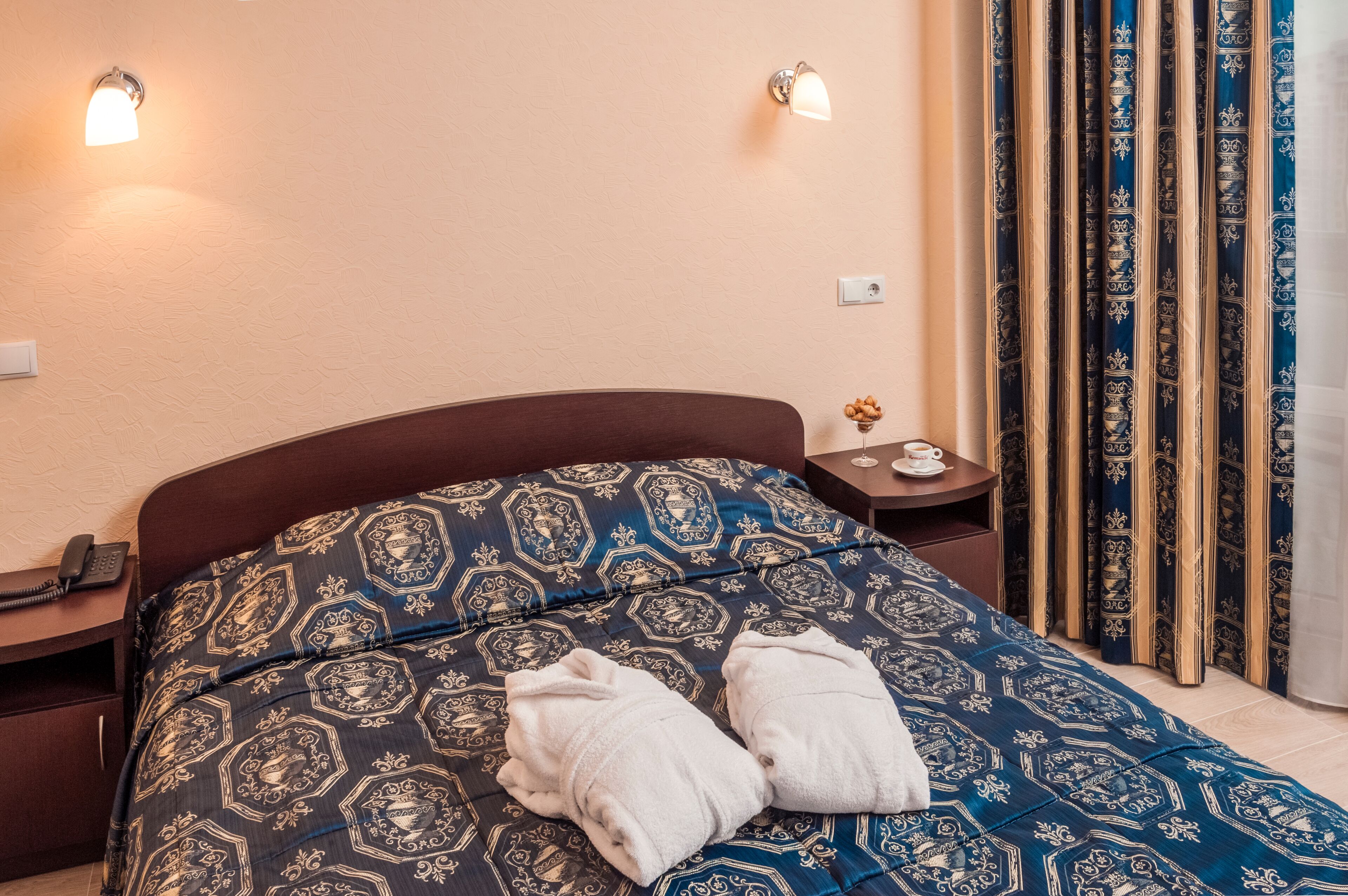 Una cama bien hecha con una colcha estampada, dos toallas, lámparas de noche, una mesita de noche, una pequeña mesa con una taza y un adorno, y cortinas con un diseño detallado.