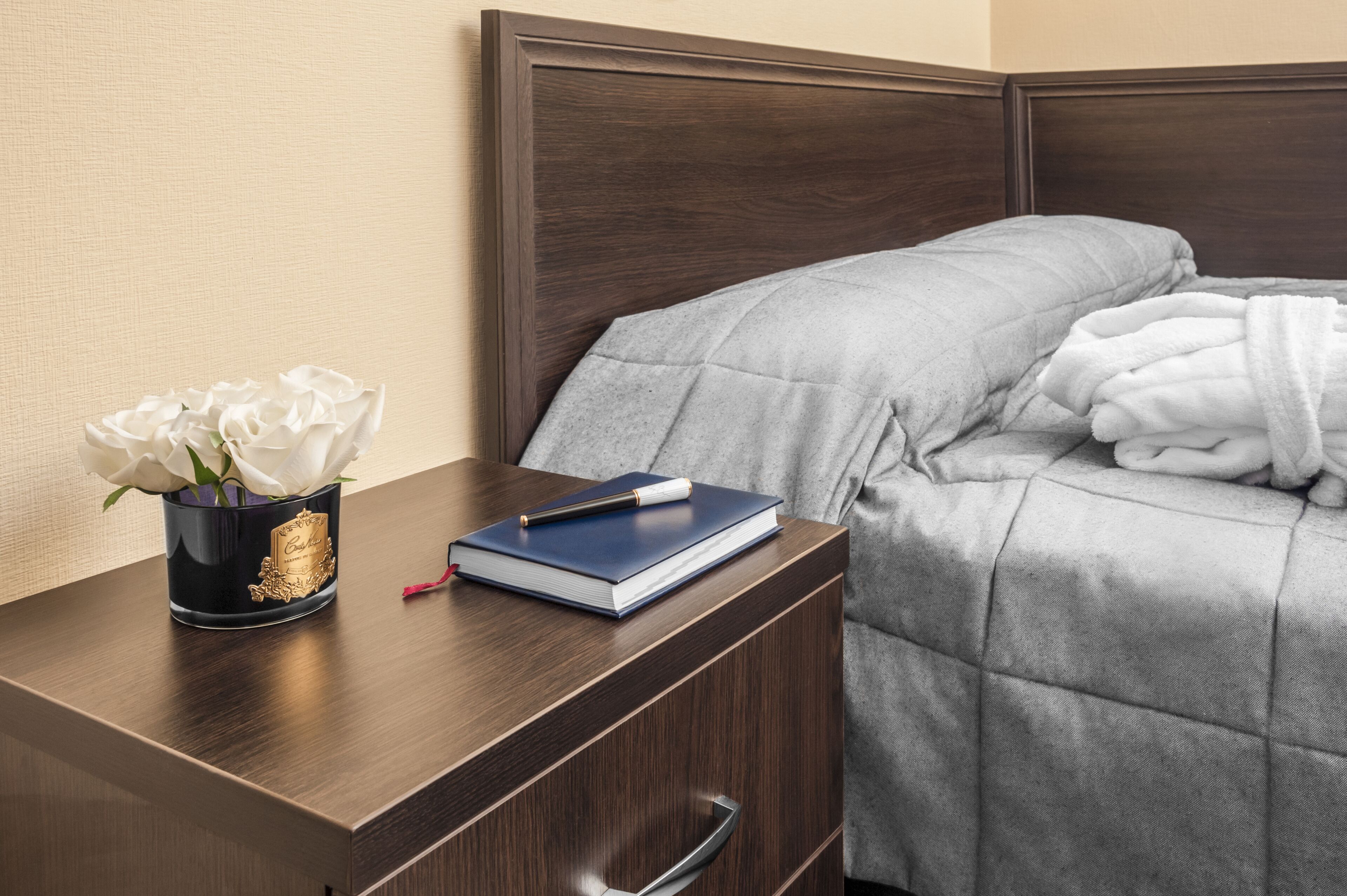 A neatly made bed with a grey blanket and a pillow, a bedside table with a book and a pen, and a vase with white flowers.