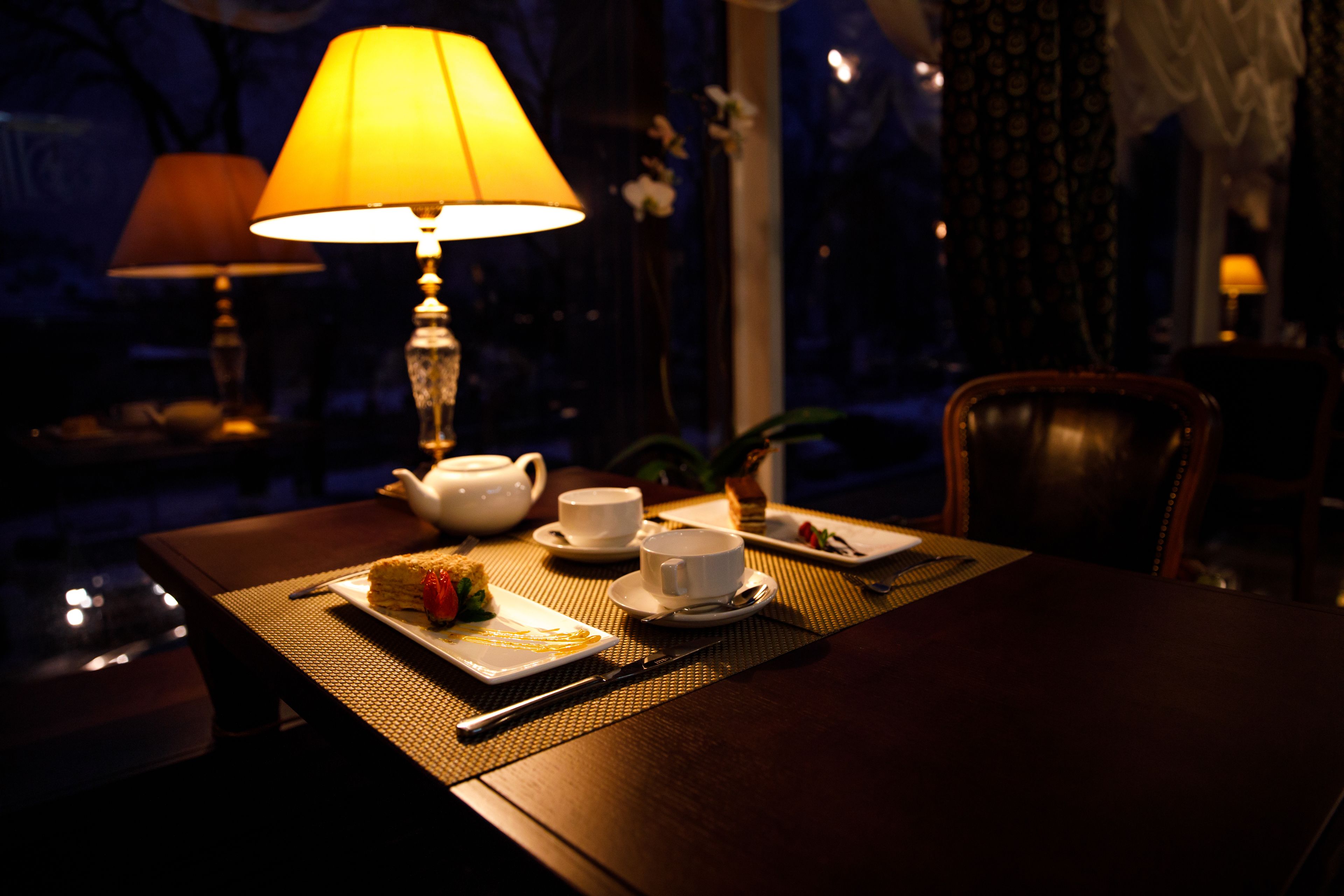 A dimly lit dining area with a table set for two, featuring a teapot, cups, a plate with a dessert, and a table lamp.