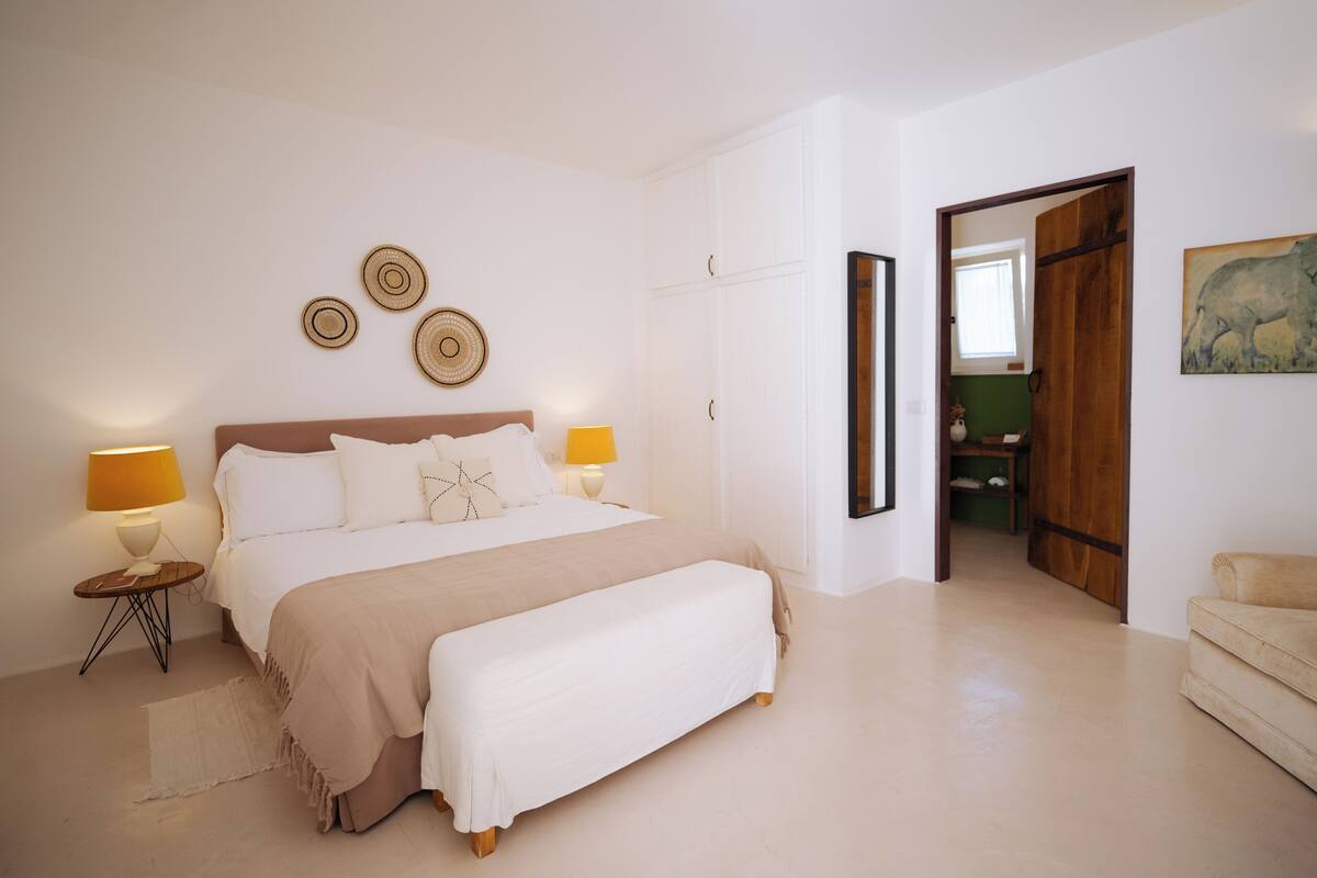 Een slaapkamer met een bed, nachtkastjes, een bankje en een deur naar een andere kamer.