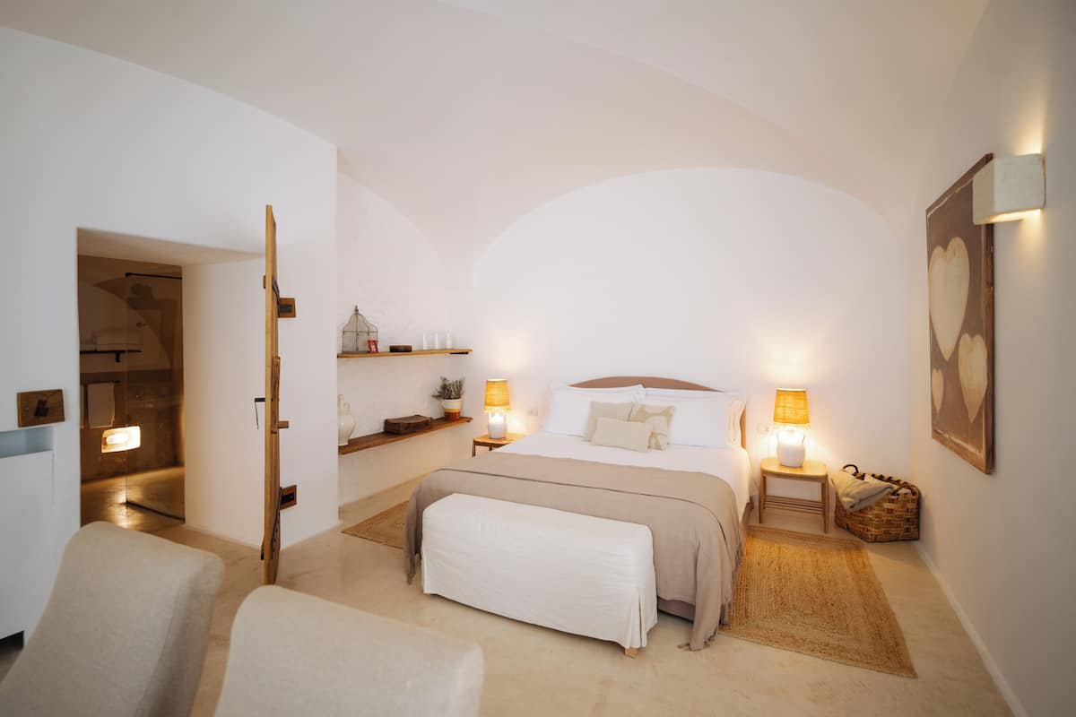 Een slaapkamer met een groot bed, bedlampen, een bankje, een klein tafeltje en een wandplank.