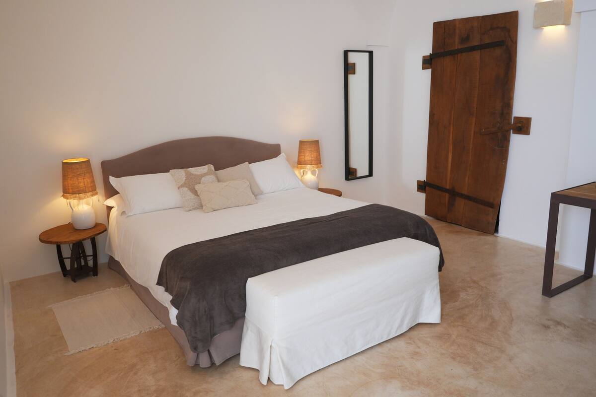 Een netjes opgemaakt bed met hoofdbord, twee bedlampen, een houten deur en een klein tafeltje ernaast.