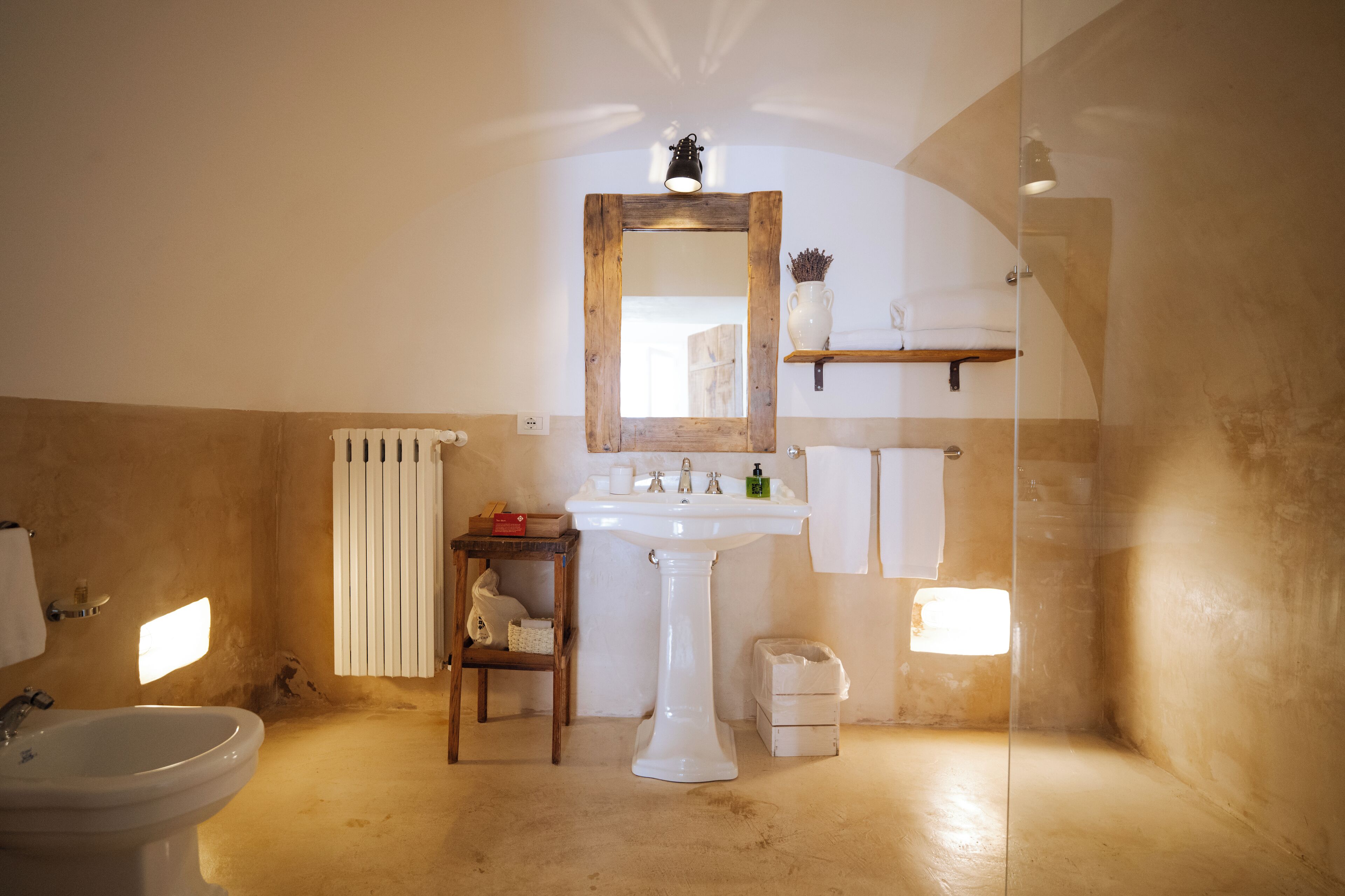 Un baño con lavamanos pedestal, un espejo, un perchero para toallas, un estante con artículos de aseo y un taburete de madera pequeño.