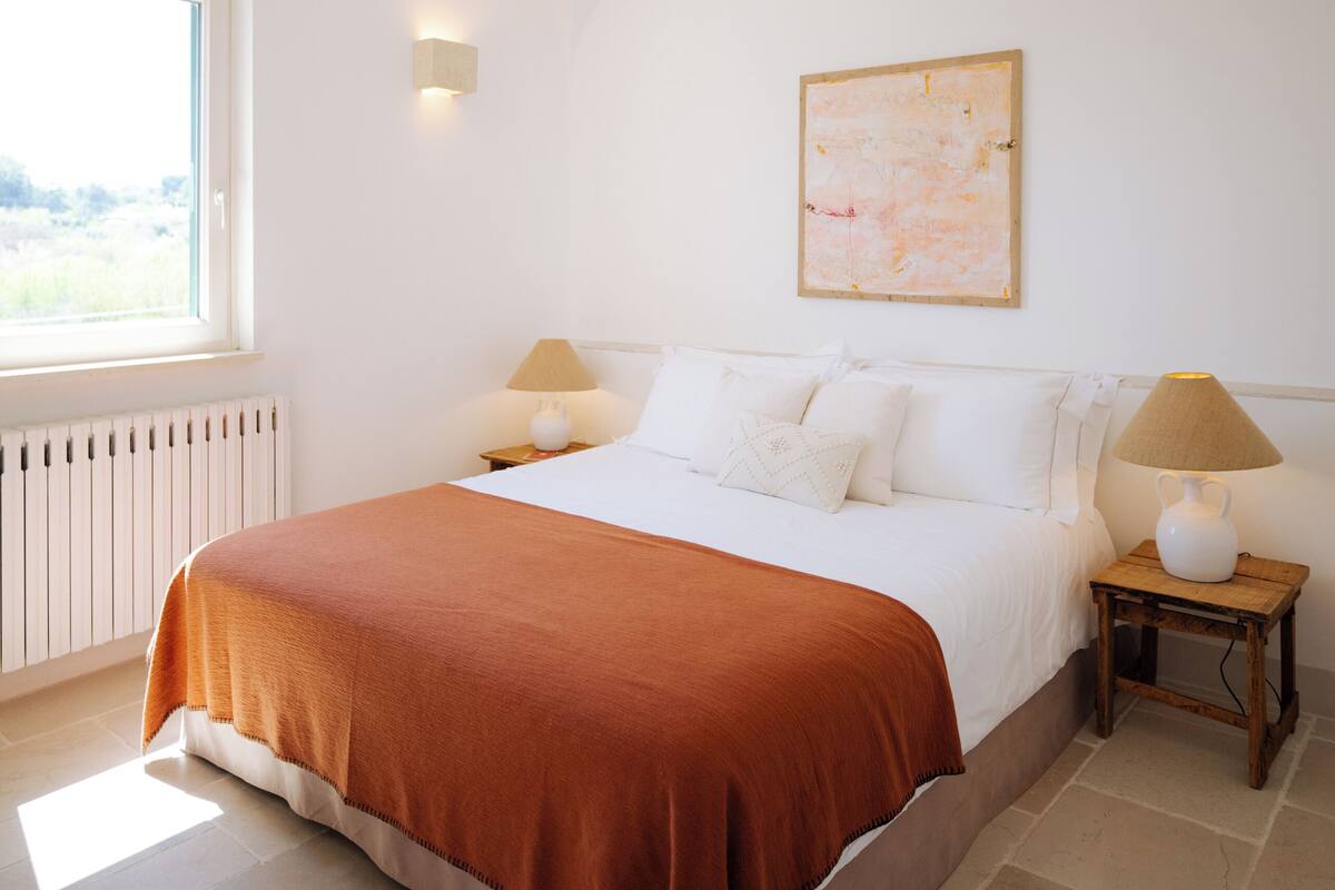 Een slaapkamer met een bed, een plafondventilator, een raam, een wandlamp en een schilderij.