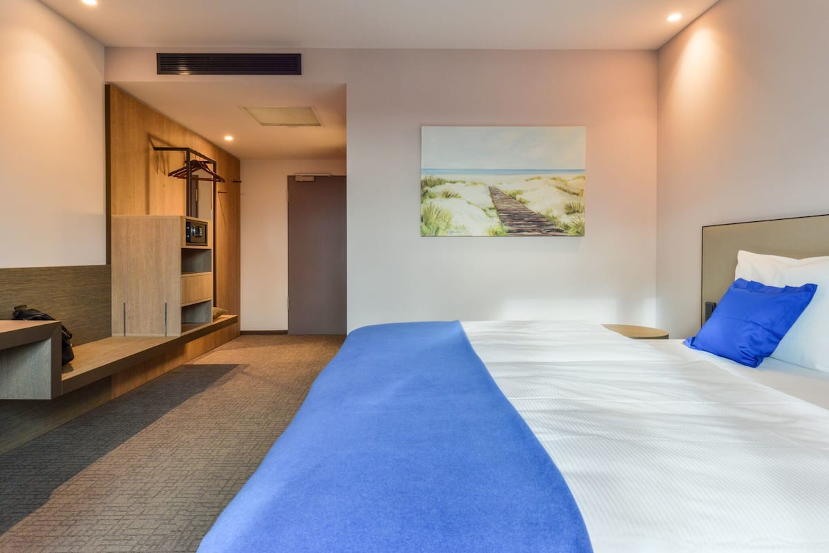 A hotel room with a bed, a blue bedspread, a painting of a beach, a wooden wardrobe, and a small desk with a chair.