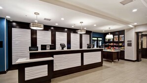 Una moderna zona de recepción de hotel con mostrador, quioscos de registro digital y una pequeña selección de aperitivos y bebidas.