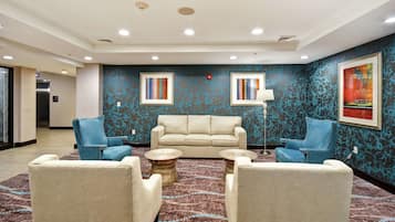 Um hall com um sofá bege, duas poltronas azuis e uma mesa de centro. Há duas obras de arte emolduradas na parede e um abajur.