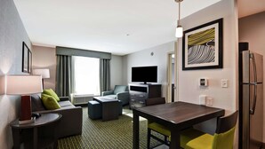 Habitación de hotel moderna con sofá, mesa de comedor, televisión de pantalla plana, refrigerador y un amplio ventanal con cortinas.
