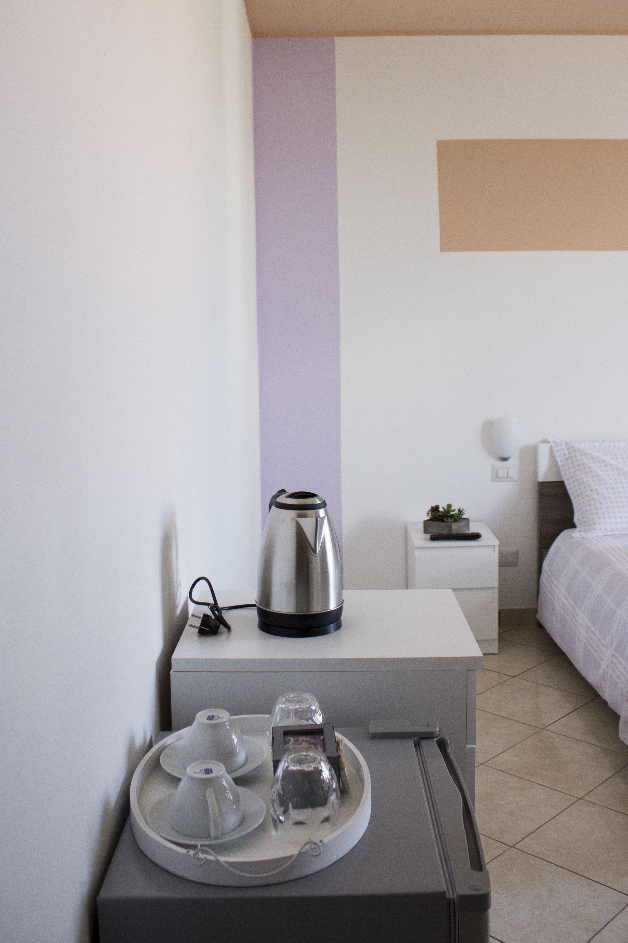 Une chambre d’hôtel bien rangée, avec un lit, une table de chevet sur laquelle se trouve une bouilloire, un plateau avec des tasses et un verre, et une petite plante posée sur la table de chevet.