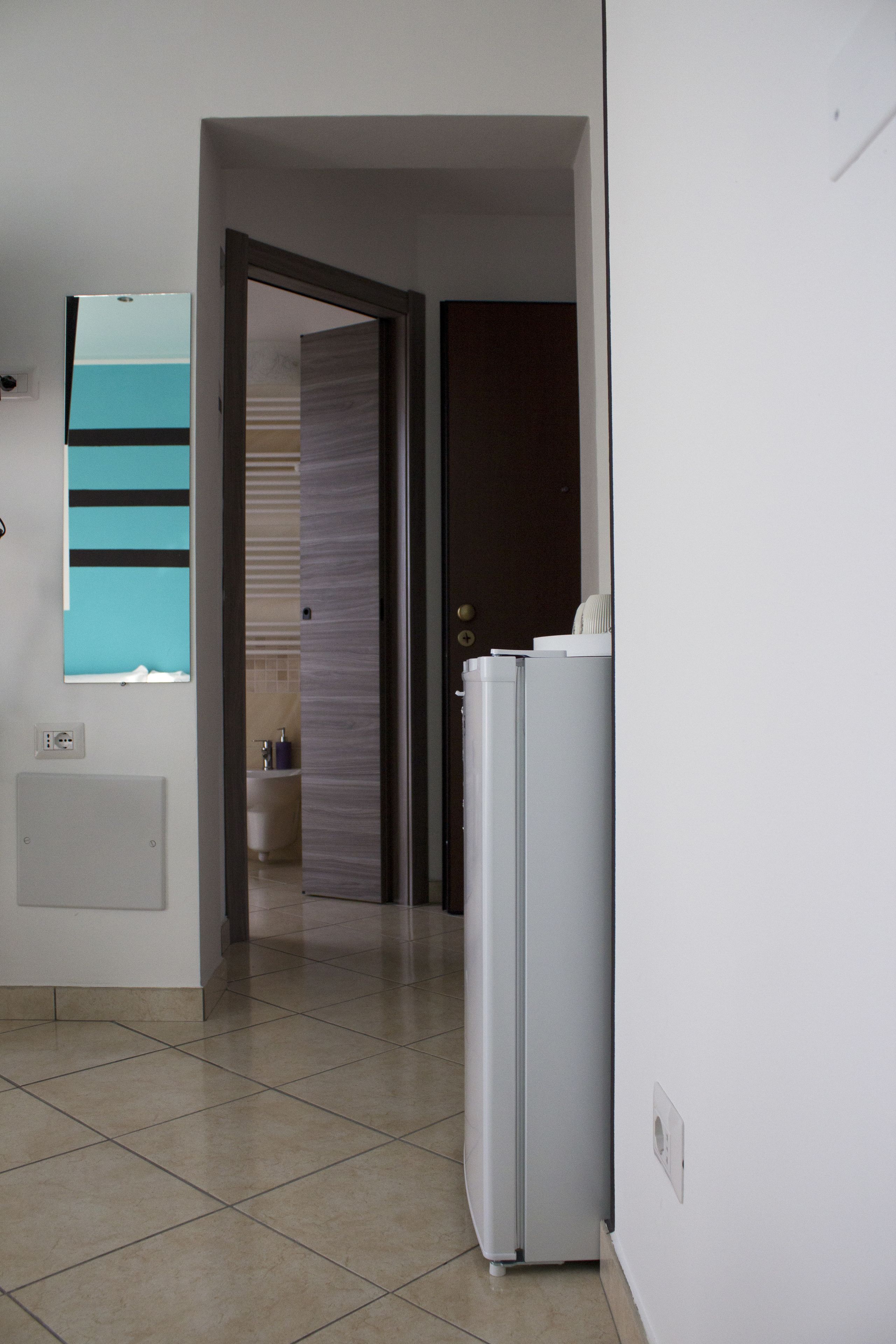 A hallway with a refrigerator, a door leading to a bathroom, and a window with blinds.