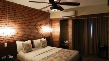 A hotel room with a brick wall, a bed with floral bedding, a ceiling fan, and an air conditioning unit.