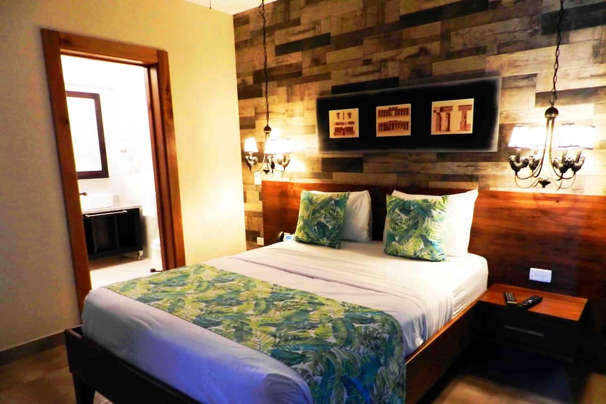 A hotel room with a bed, a wooden headboard, a nightstand, a lamp, and a wall-mounted picture frame.