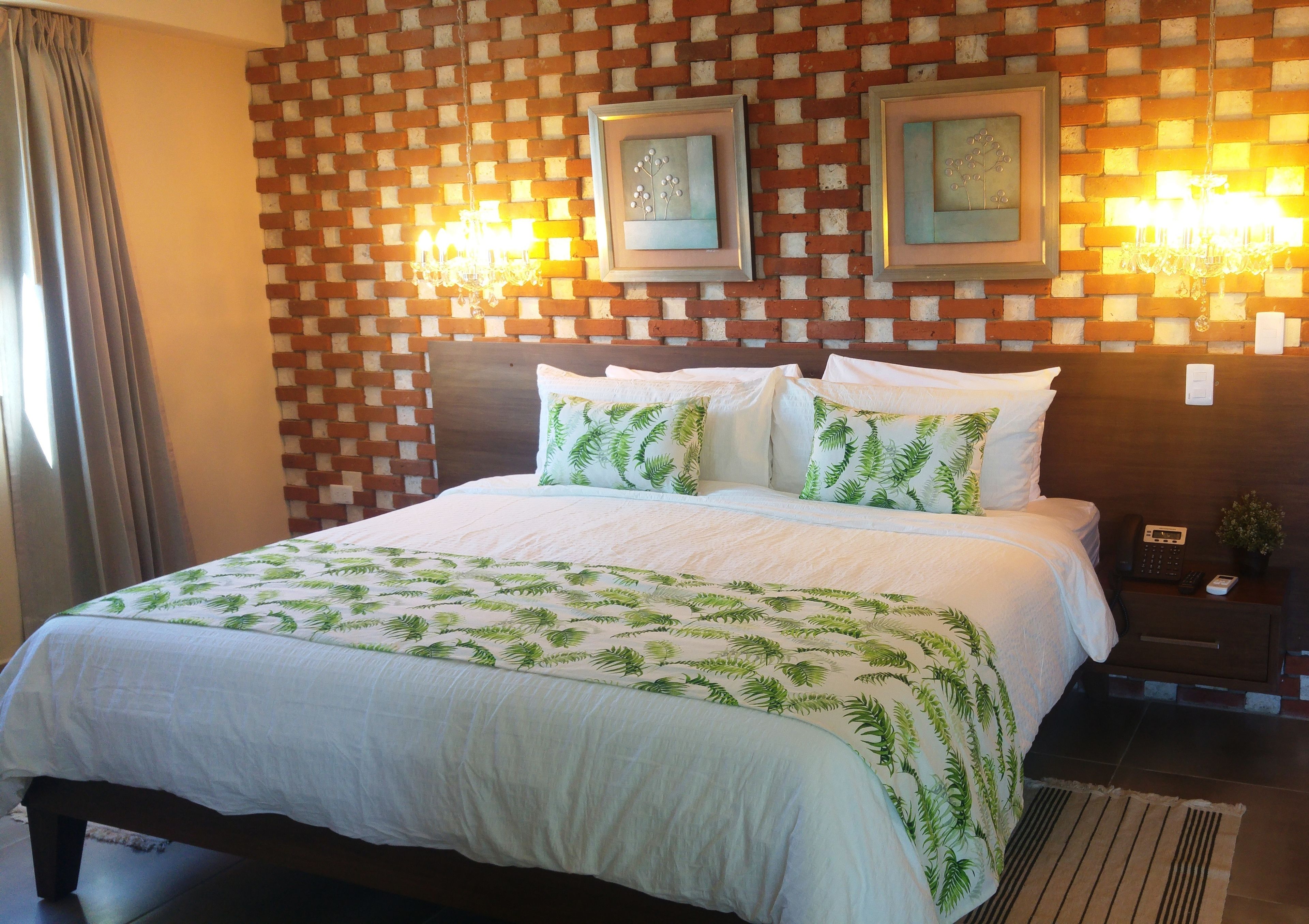 Una cama bien hecha con una colcha blanca y verde estampada, un cabecero de madera y dos lámparas fijadas a la pared.