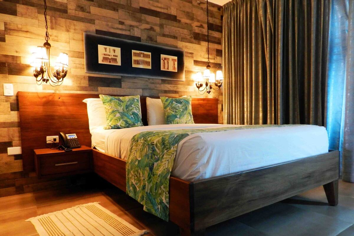 A bedroom with a wooden headboard, a bed with a green leaf-patterned blanket, two wall-mounted lamps, and a telephone on a bedside table.