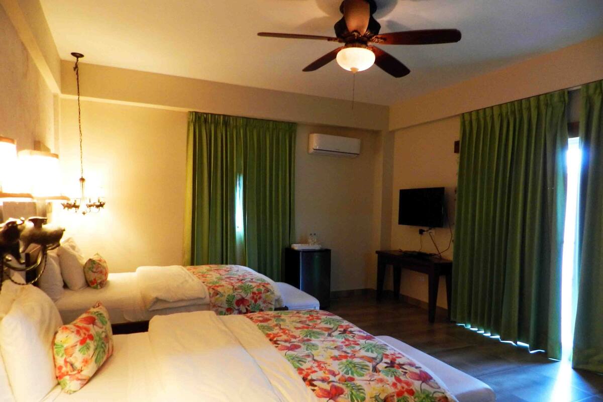 A hotel room with two beds, a ceiling fan, an air conditioning unit, a television, and green curtains.