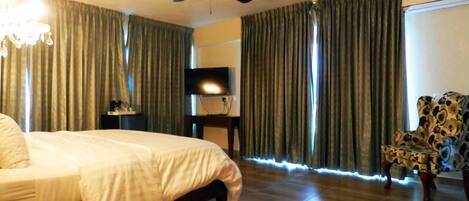 Una habitación de hotel con una cama, ventilador de techo, televisión, silla y cortinas.