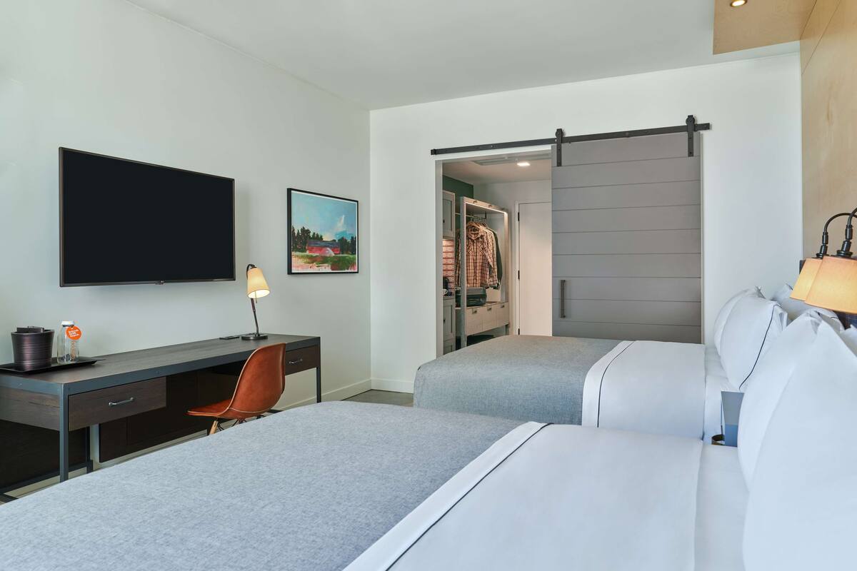 A modern hotel room with a flat-screen TV, a desk with a lamp, two beds with grey bedding, a closet with sliding doors, and a painting on the wall.