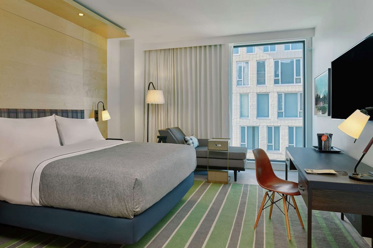 A hotel room with a bed, desk, chair, and a large window with a city view.