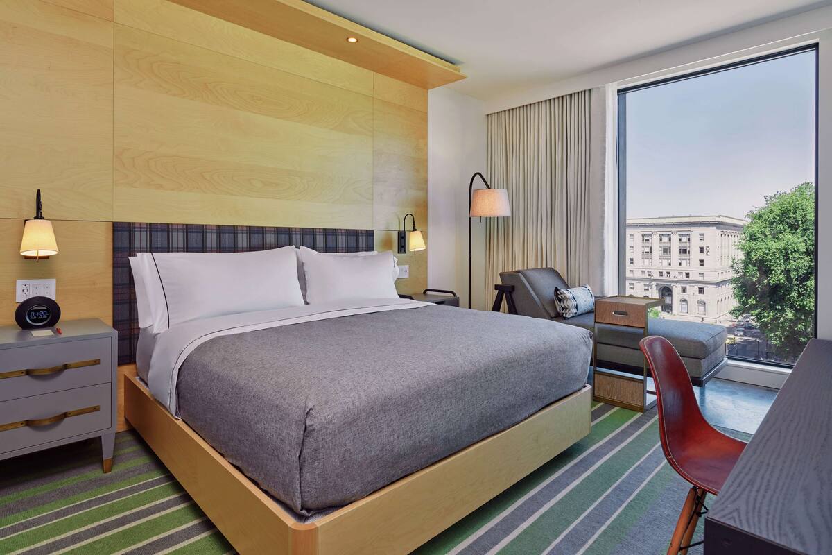 A hotel room with a large bed, bedside lamps, a desk with a chair, a view of a building through a window, and a cityscape.