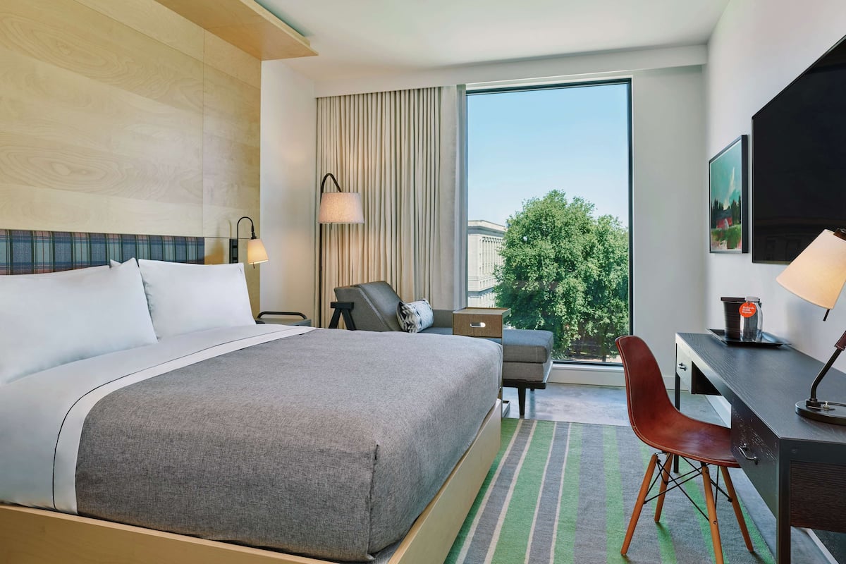 A hotel room with a large bed, a desk, a chair, and a view of a tree through a window.