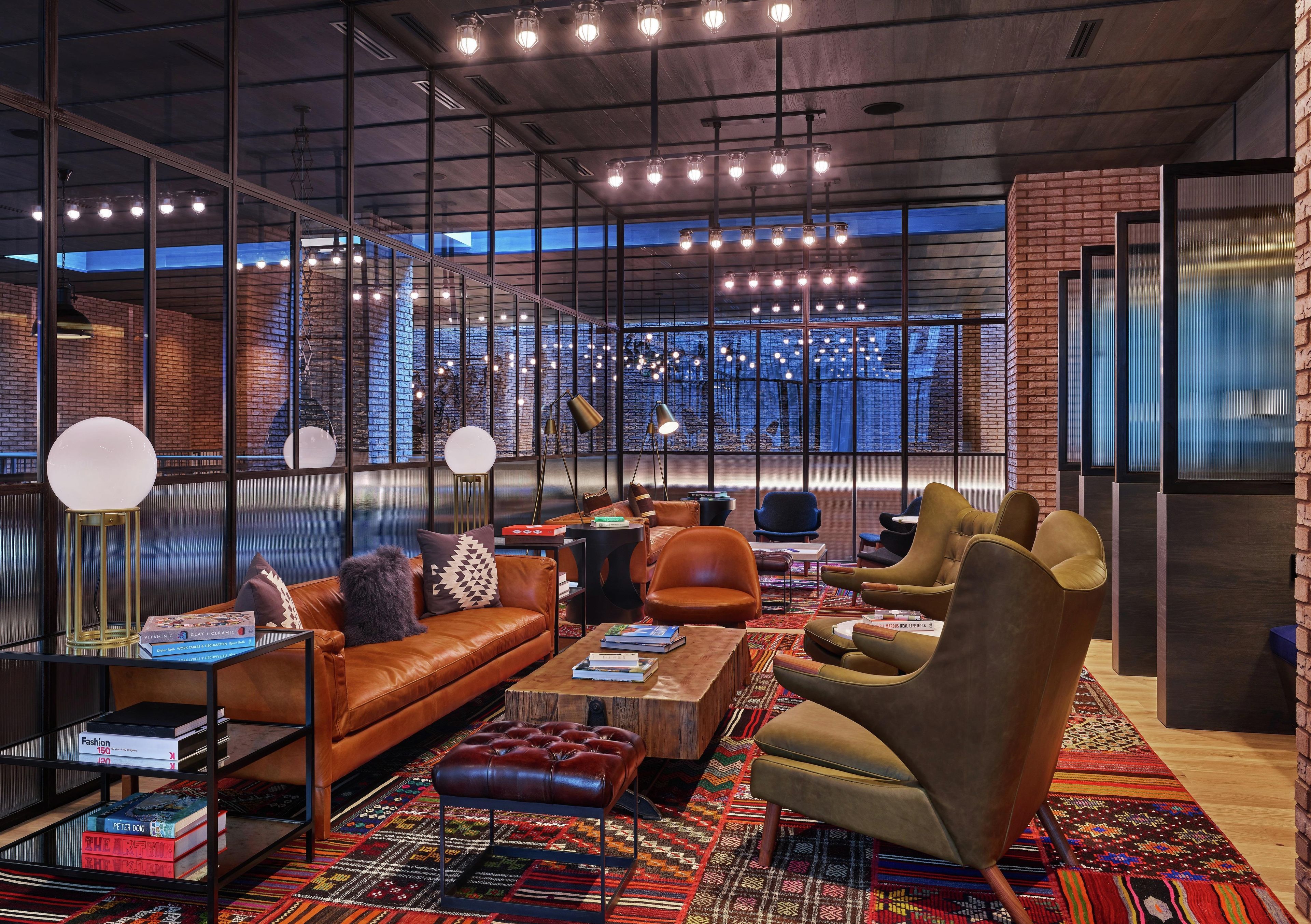 A modern lounge area with leather sofas, armchairs, and a coffee table. There are books and a lamp on a side table. The room has large windows with a view of the cityscape.