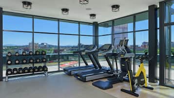 A gym with treadmills, dumbbells, and exercise bikes, large windows with a city view.