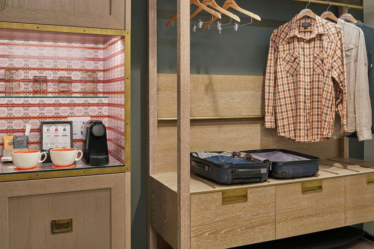 A hotel room with a wooden wardrobe, a glass display cabinet with cups and a coffee maker, and a hanging wardrobe with clothes and a suitcase.