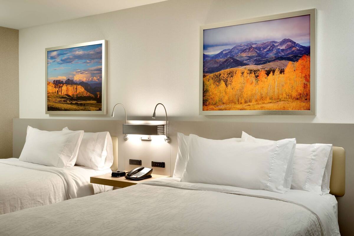 A hotel room with two beds, a bedside table with a phone, and two framed landscape paintings on the walls.