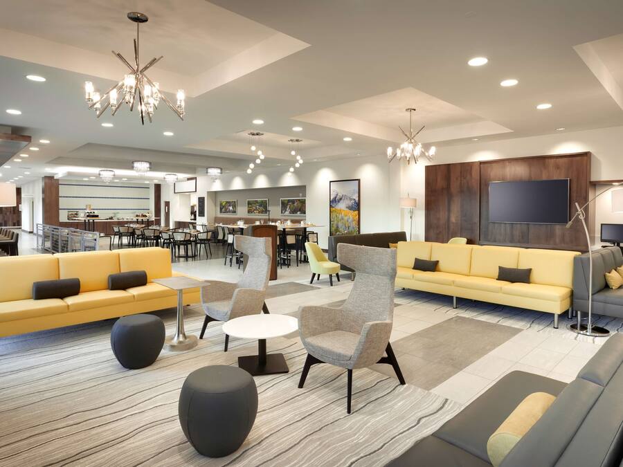 A spacious hotel lobby with modern furnishings, a dining area, and a large flat-screen TV mounted on the wall.