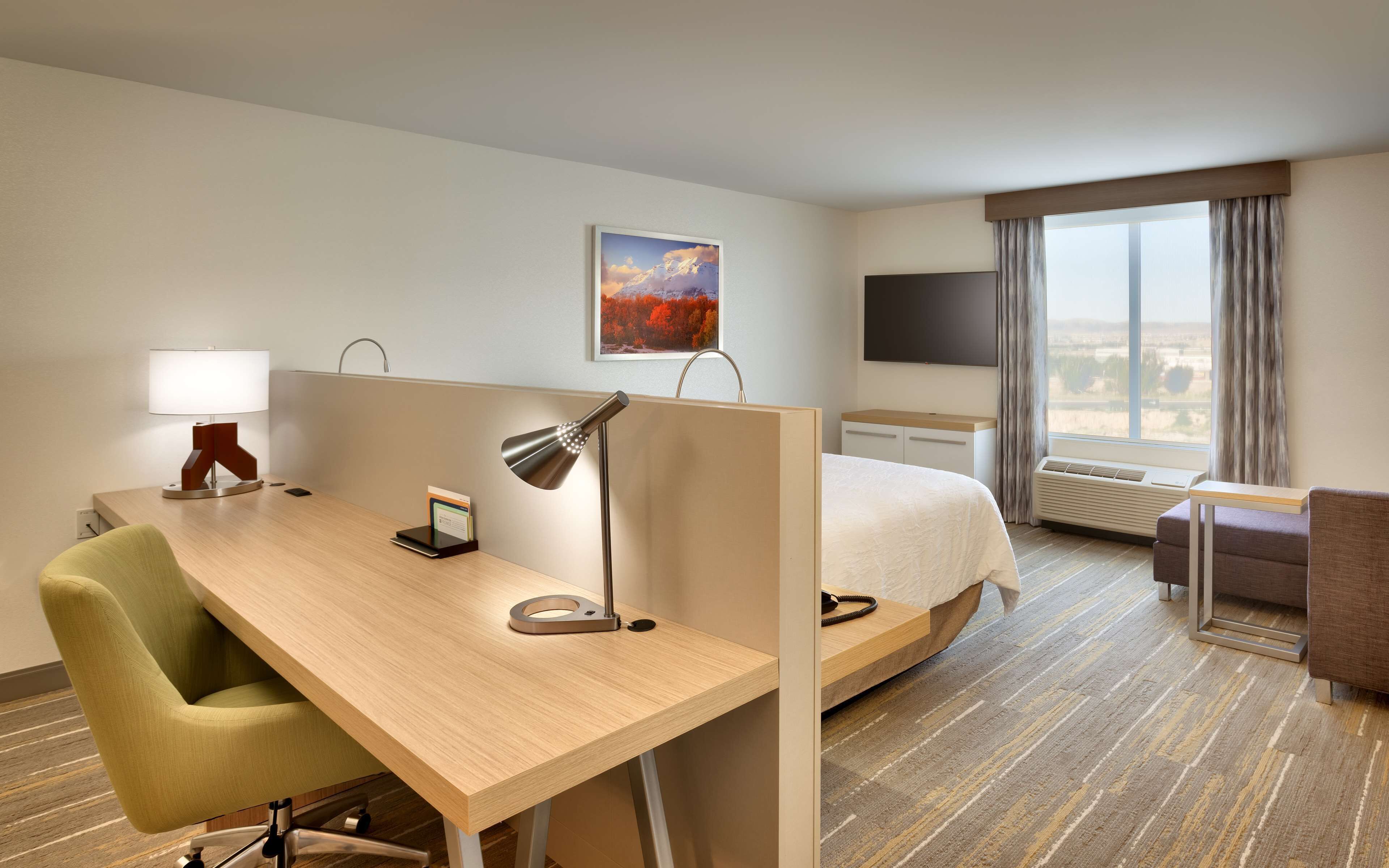 Une chambre d’hôtel avec un bureau en bois, une chaise verte, un lit, une télévision et une fenêtre avec des rideaux.