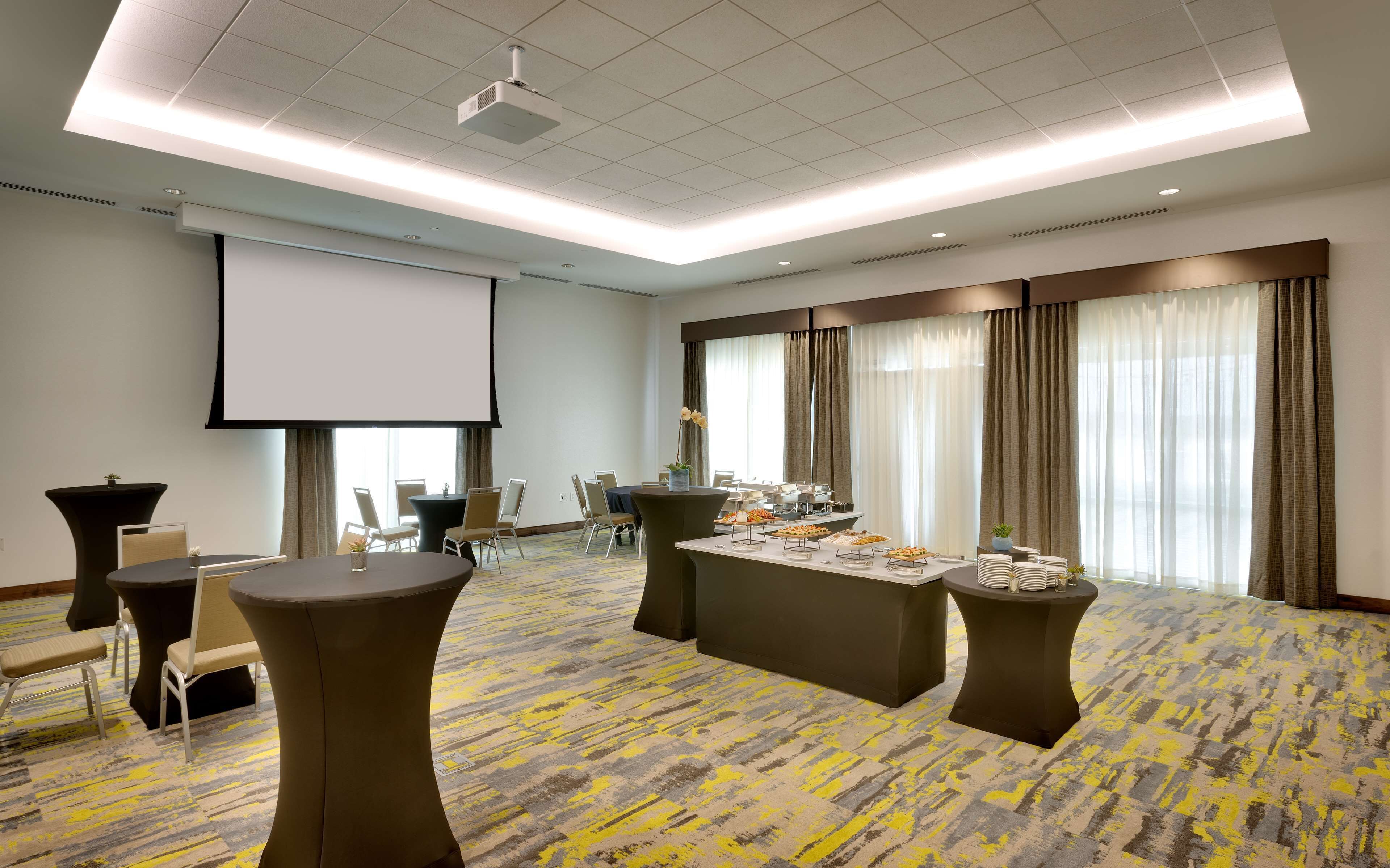 Un salón de conferencias con una pantalla grande, varias mesas con sillas y piso alfombrado.