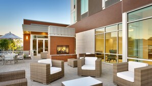 A patio area with wicker furniture and a fireplace, overlooking a building with large windows. - Hilton Garden Inn Lehi (Lehi)