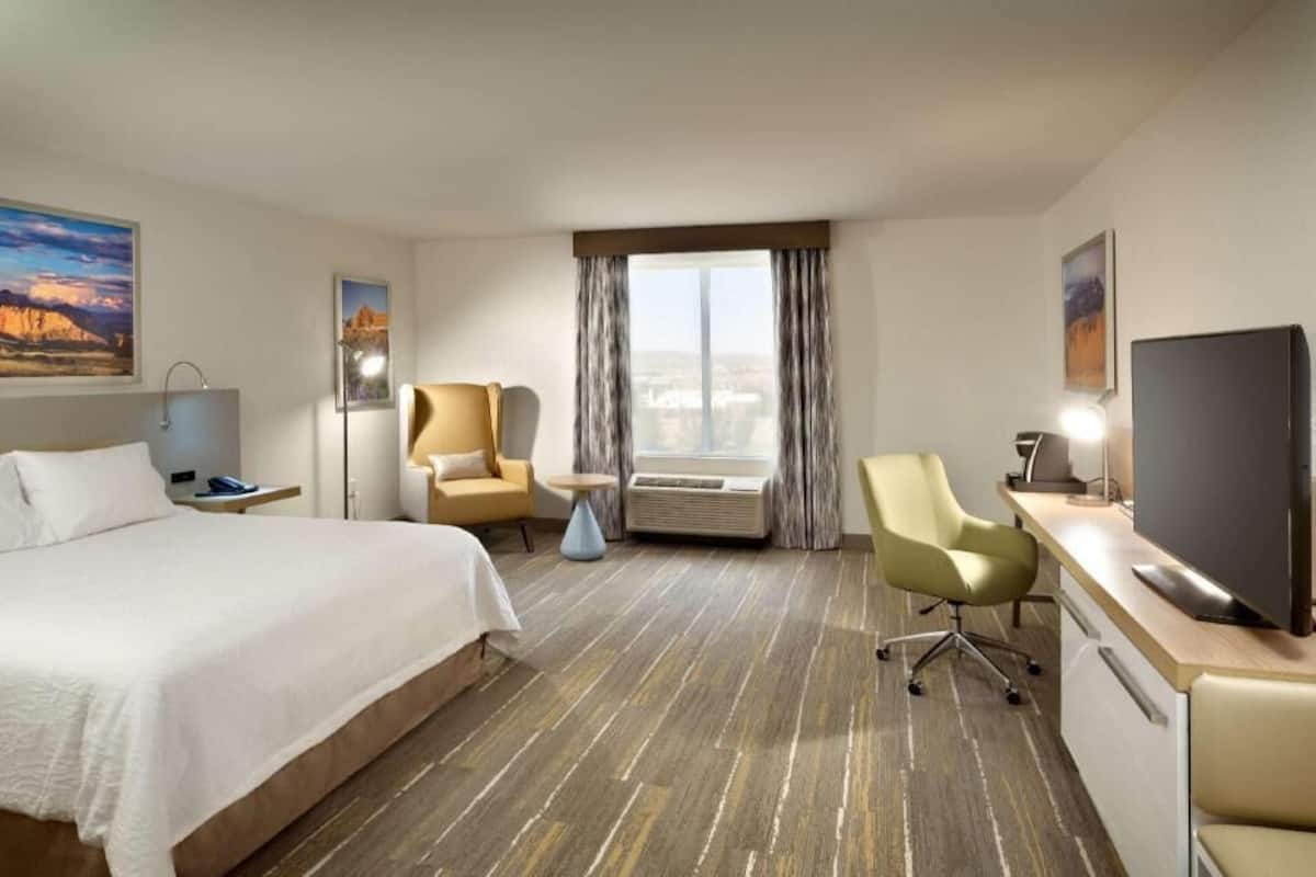 A hotel room with a bed, a desk with a flat-screen TV, a chair, a small table, and a window with curtains.