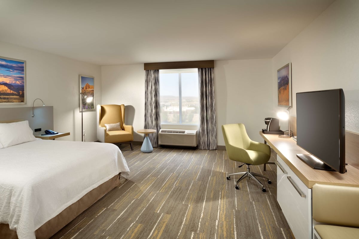 A hotel room with a bed, a desk with a flat-screen TV, a chair, an air conditioner, and a large window with curtains.